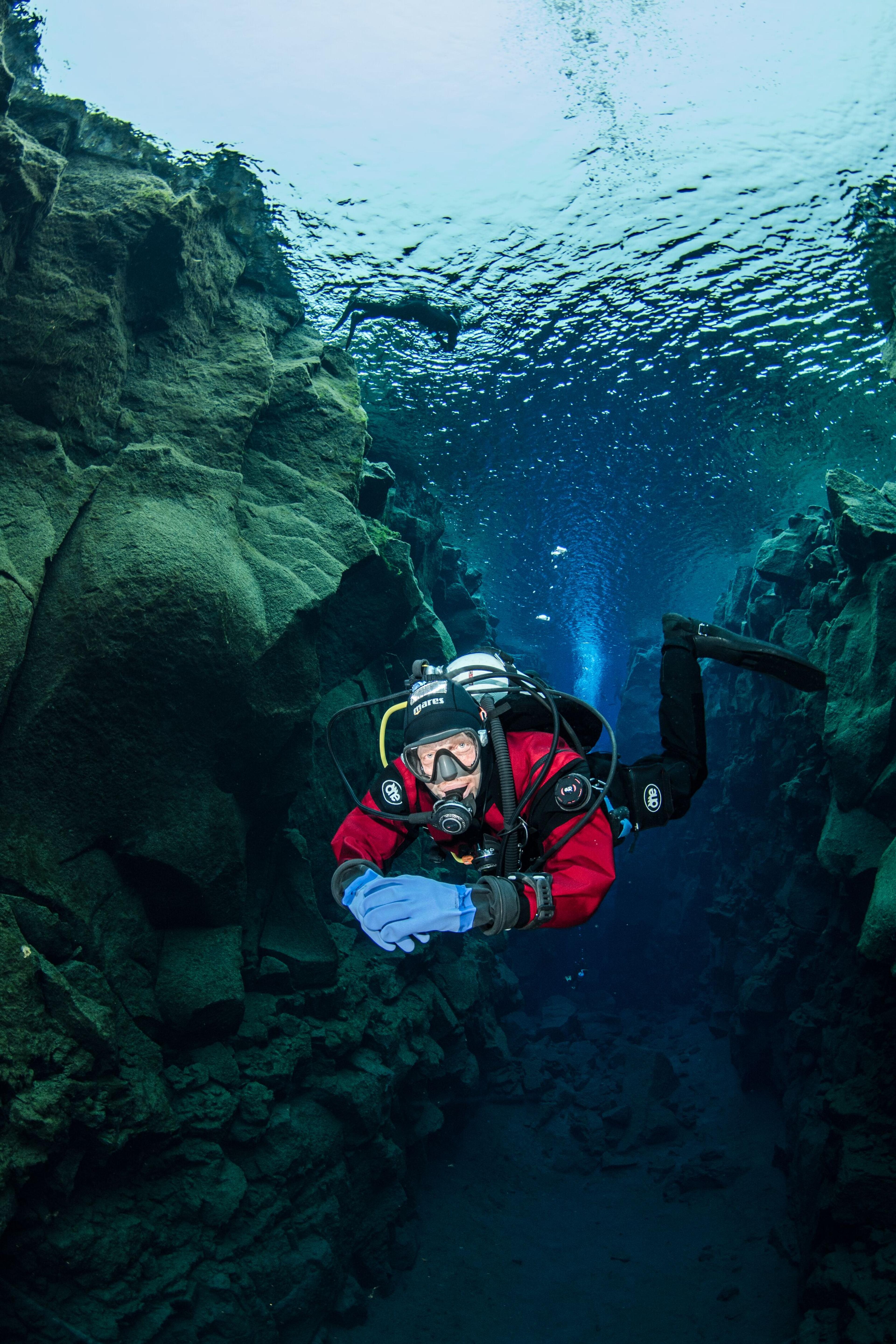 A diver in a red suit swims through a clear underwater canyon with rock walls.