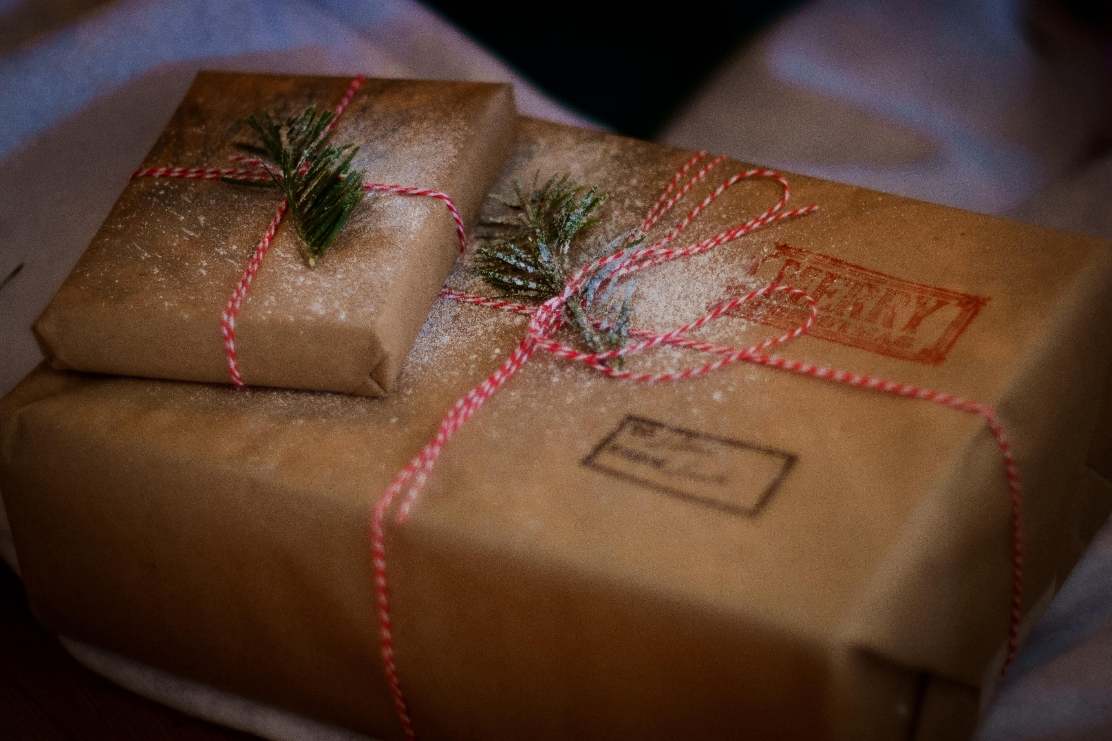 Two gifts wrapped in brown paper and tied with red-and-white string, each adorned with a small green sprig, suggesting a festive holiday theme.