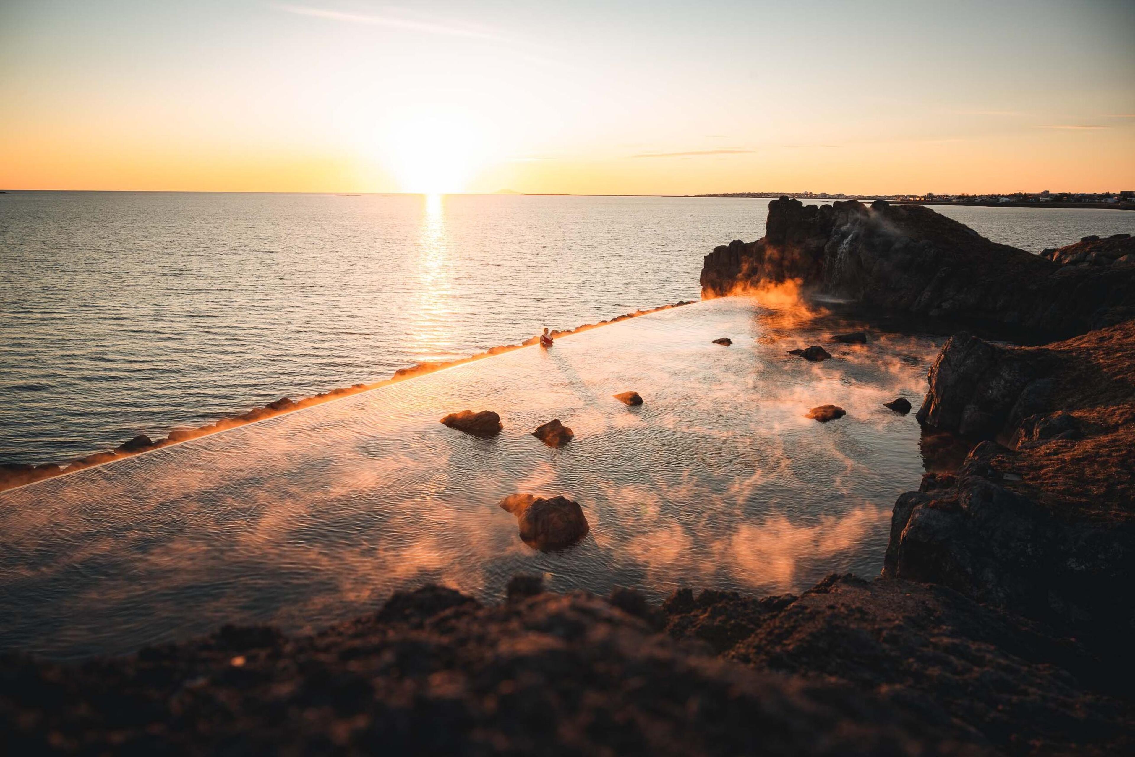 The golden hour at Sky Lagoon, where the setting sun meets the horizon, casting a warm glow over the misty geothermal waters and the rocky coast.