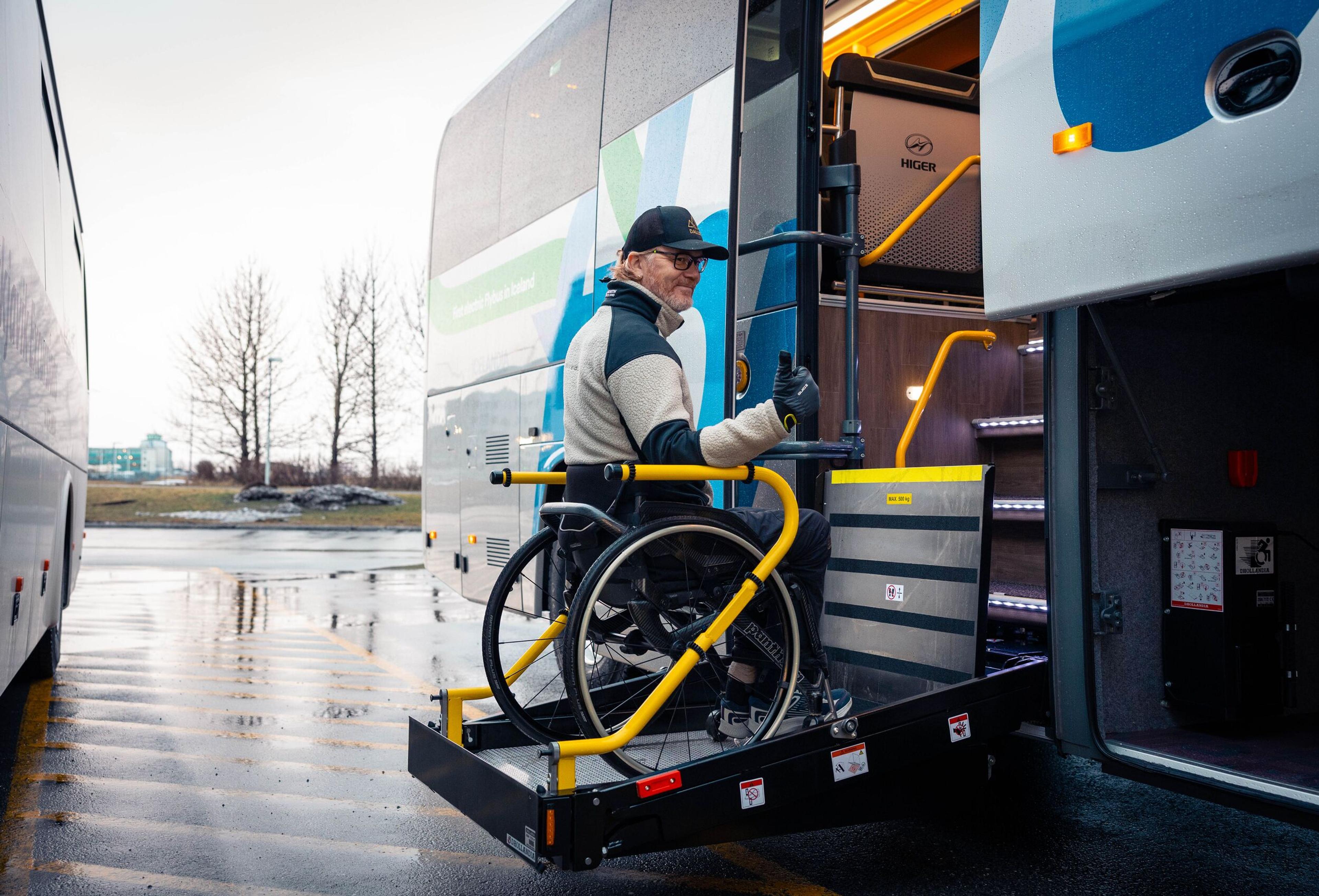A man in a wheelchair uses a hydraulic lift with yellow railings to board a bus.