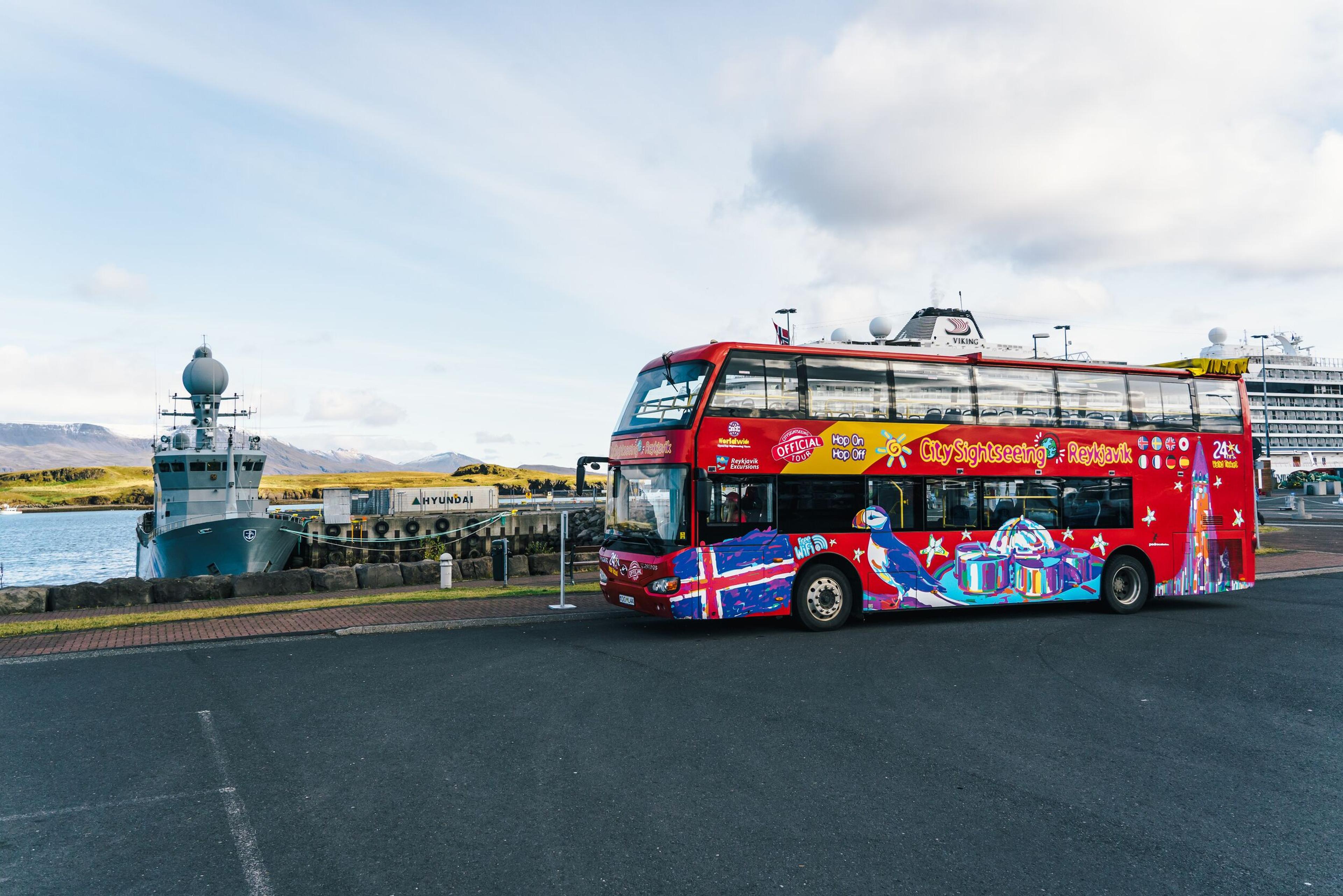 Colorful sightseeing bus parked by Reykjavik harbor, with ships and distant mountains in the background.