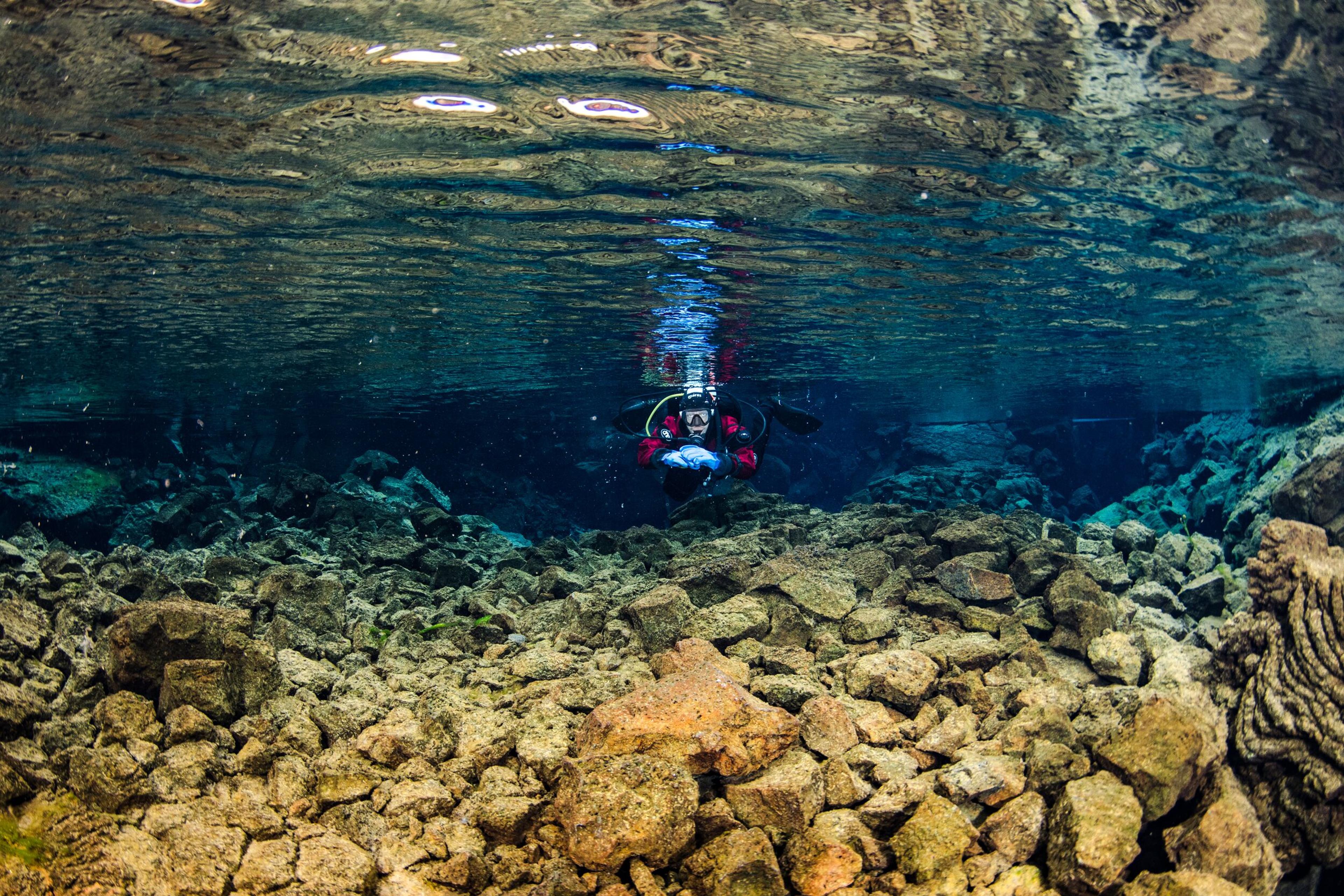 A diver in a red suit floats in clear blue water above a rocky underwater chasm.