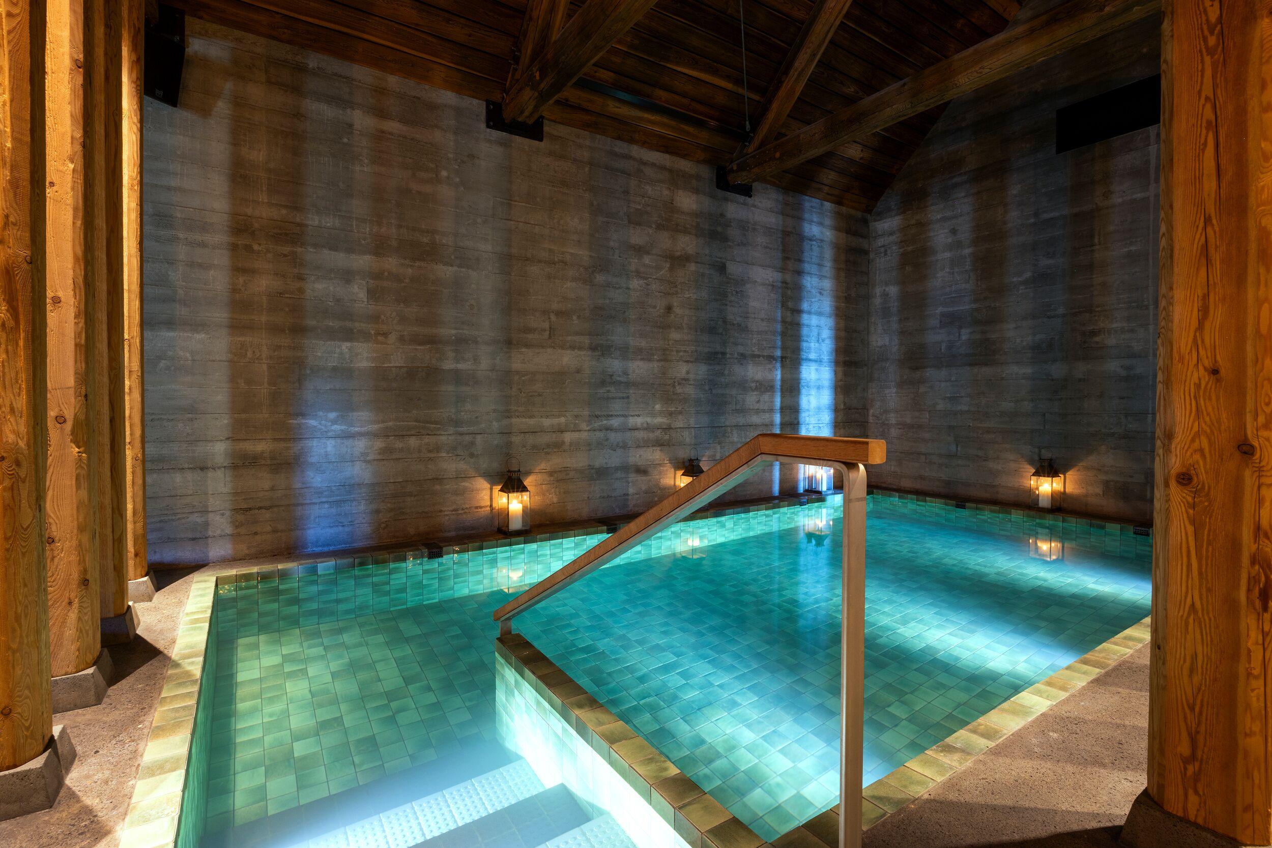 An indoor swimming pool with illuminated teal tiles, concrete walls, and a wooden beamed ceiling.