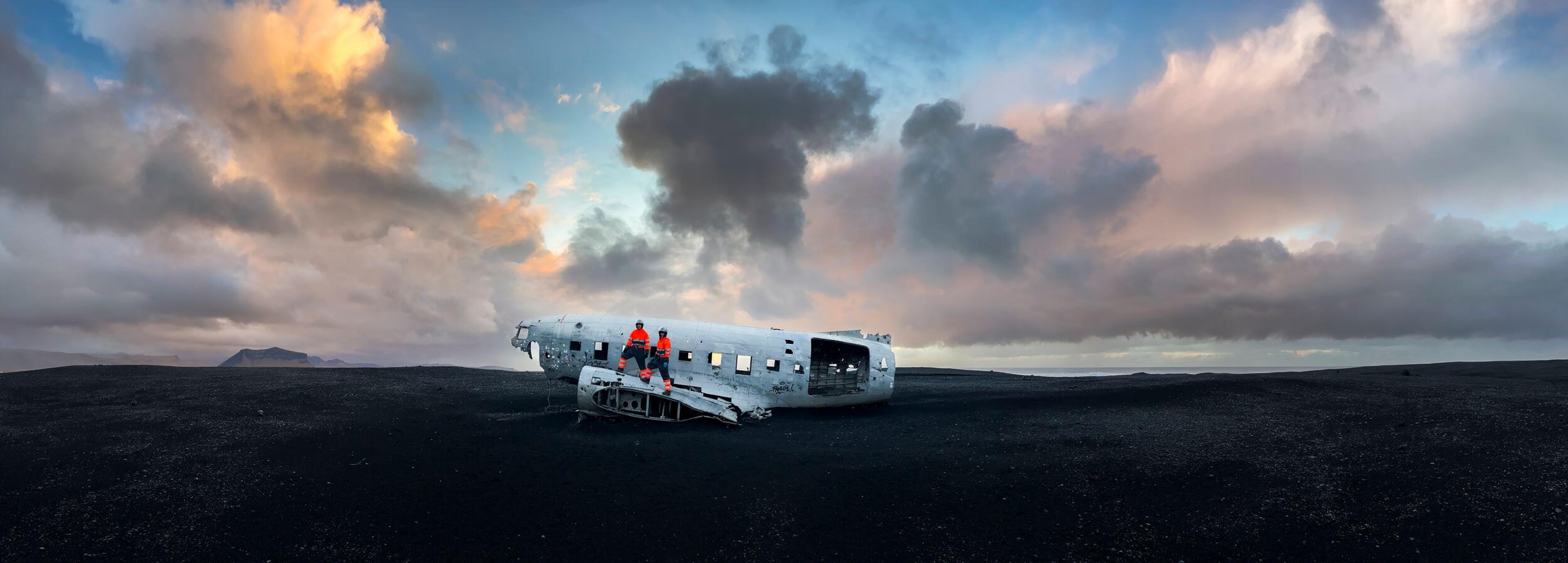 Two individuals standing on the wing of a plane wreck, with a mountain visible to the left and expansive black sand surrounding the rest.