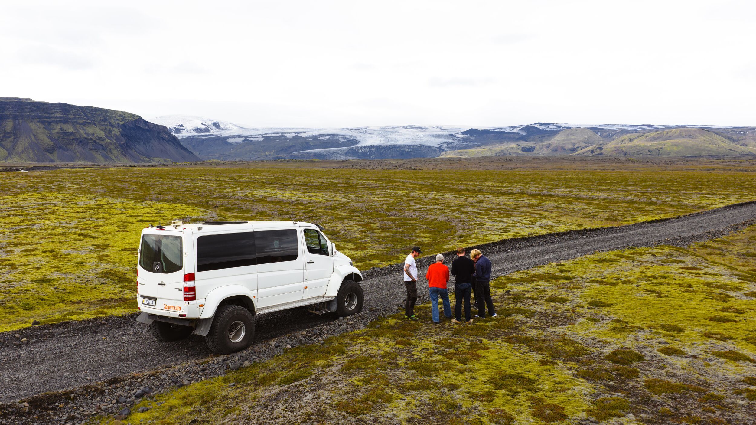 White 4x4 van and four people on a gravel road in a vast mossy landscape with mountains and a glacier.