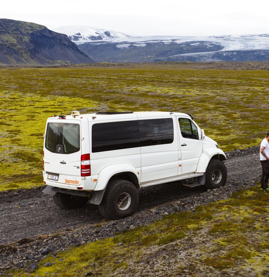 White 4x4 van and four people on a gravel road in a vast mossy landscape with mountains and a glacier.