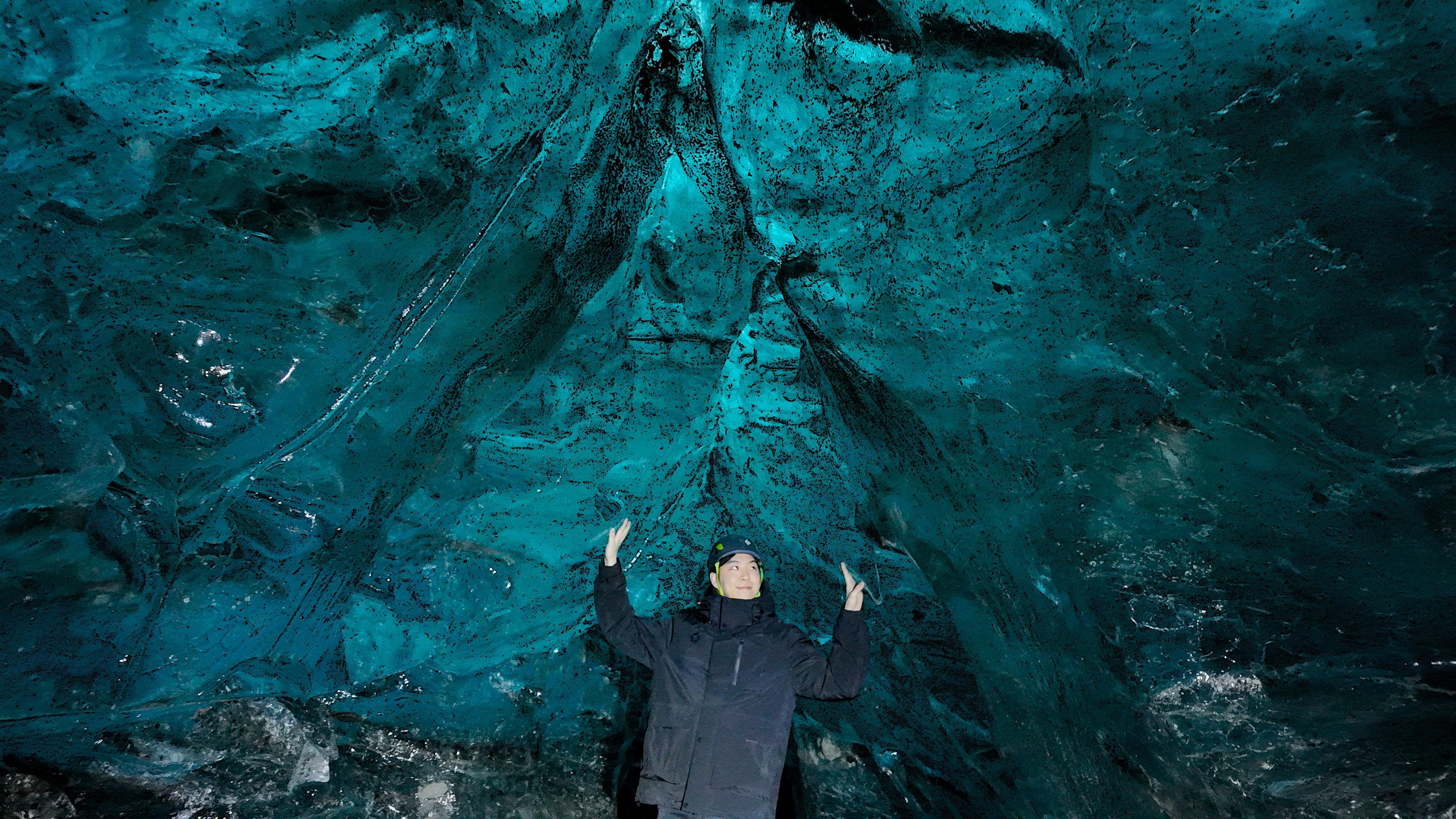 Person standing inside a vibrant blue ice cave with arms raised, looking up at the illuminated ice ceiling.