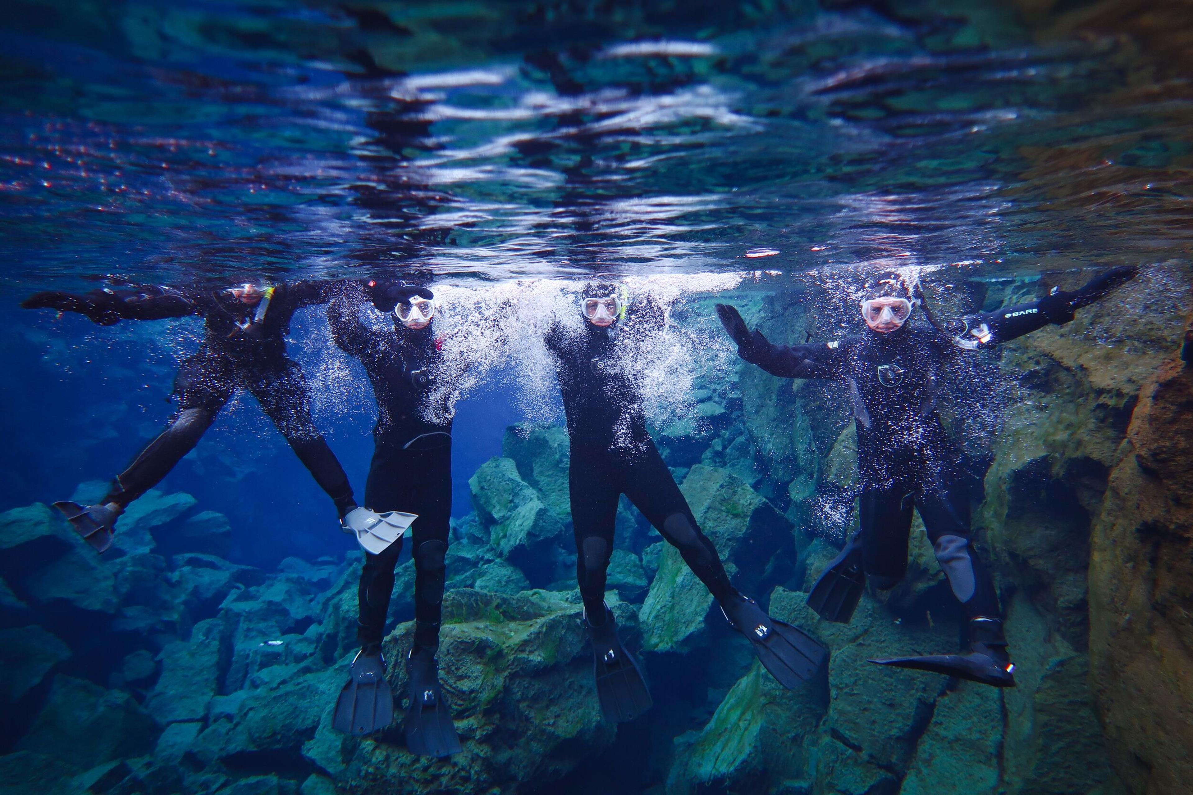 Four people in wetsuits and masks submerged in clear blue water with a rocky bottom.