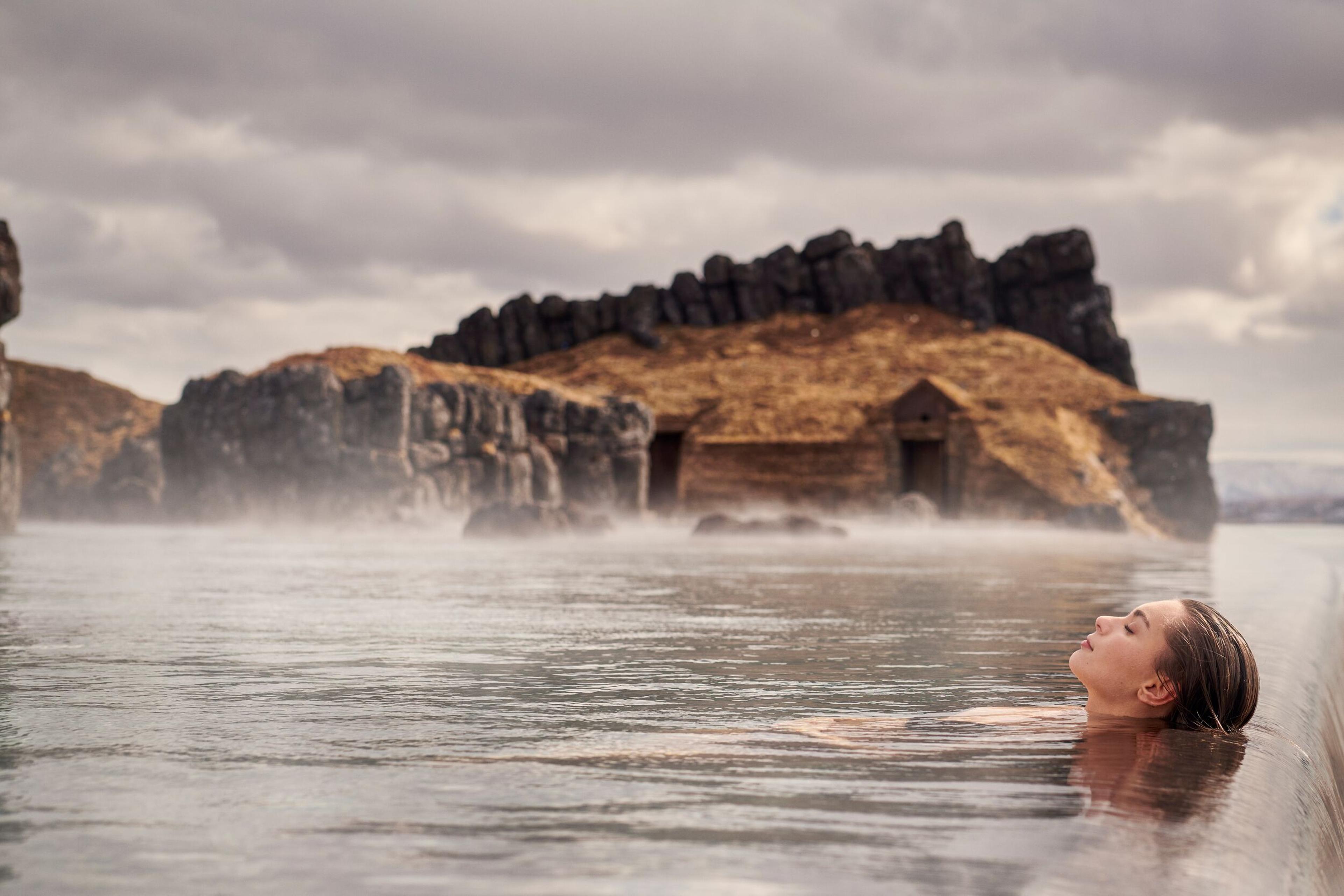 A woman immersed in the geothermal waters of Sky Lagoon Iceland, surrounded by rocky cliffs and enveloped in the ethereal mist of the warm water, under a soft, hazy sky.