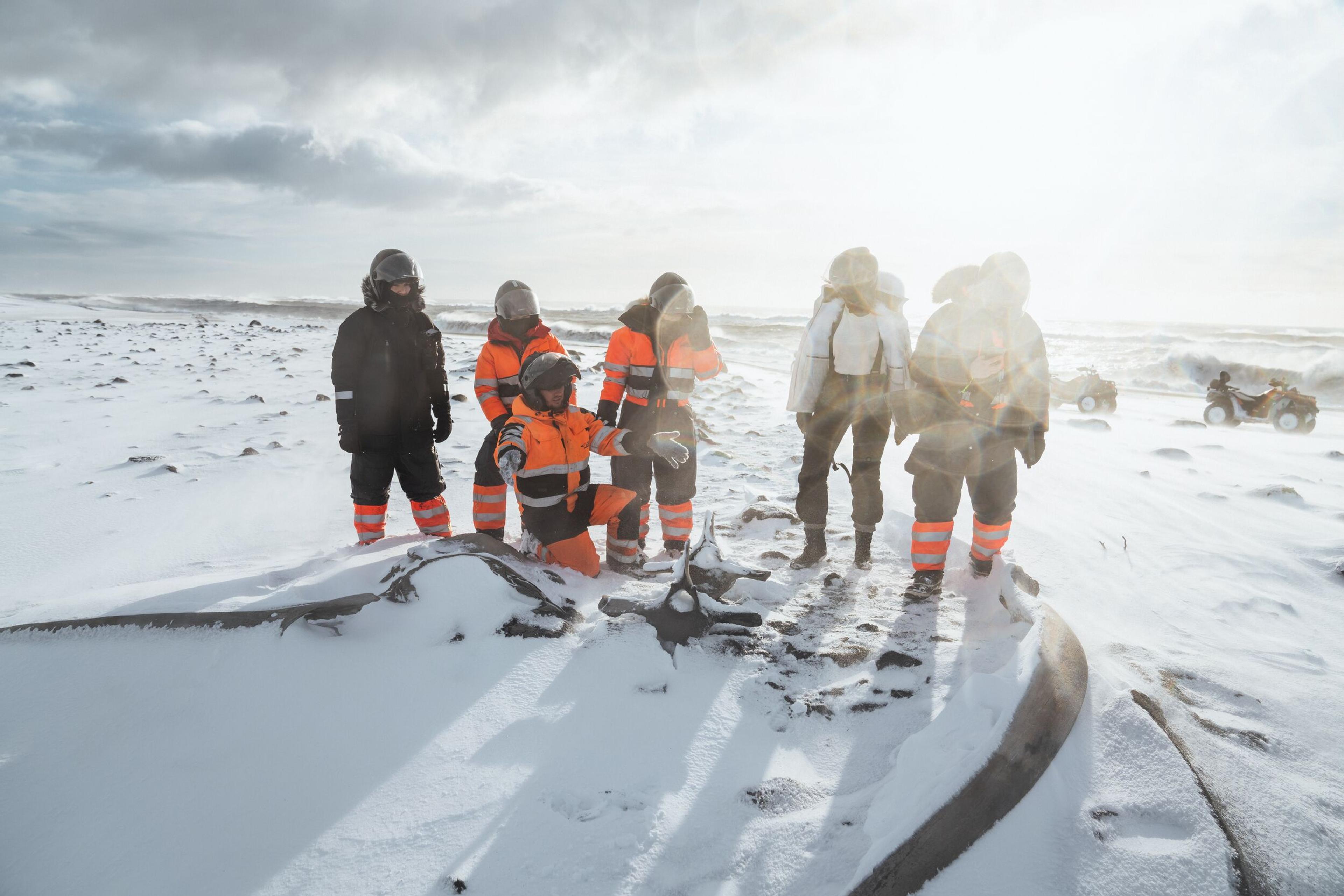 A group of adventurers in bright orange and black winter gear examine the remains of a snow-covered plane wreckage, with sunlight casting a dramatic glow over the frozen landscape.