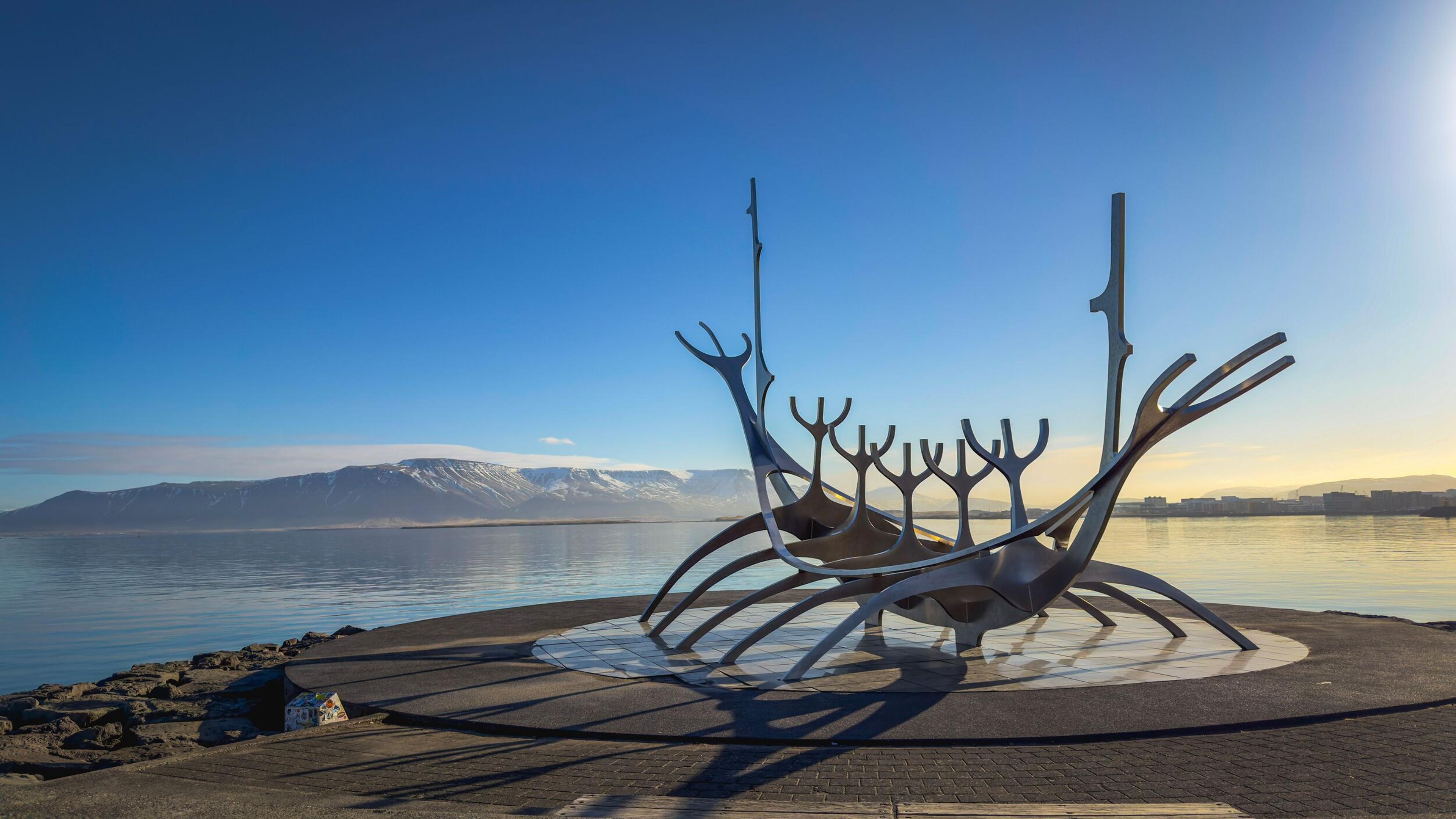 Skeletal metal sculpture of a Viking ship by a bay with snow-capped mountains under a clear blue sky.