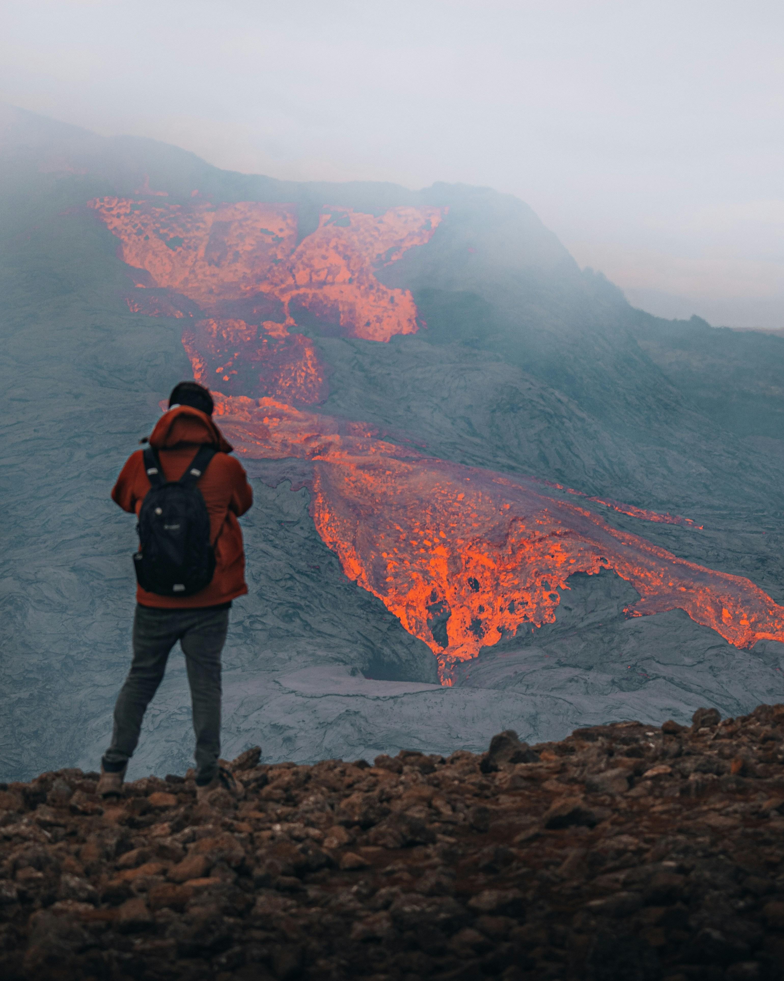 Person standing on rocky terrain observing an active volcano with glowing lava flow, highlighting the awe of nature's power.