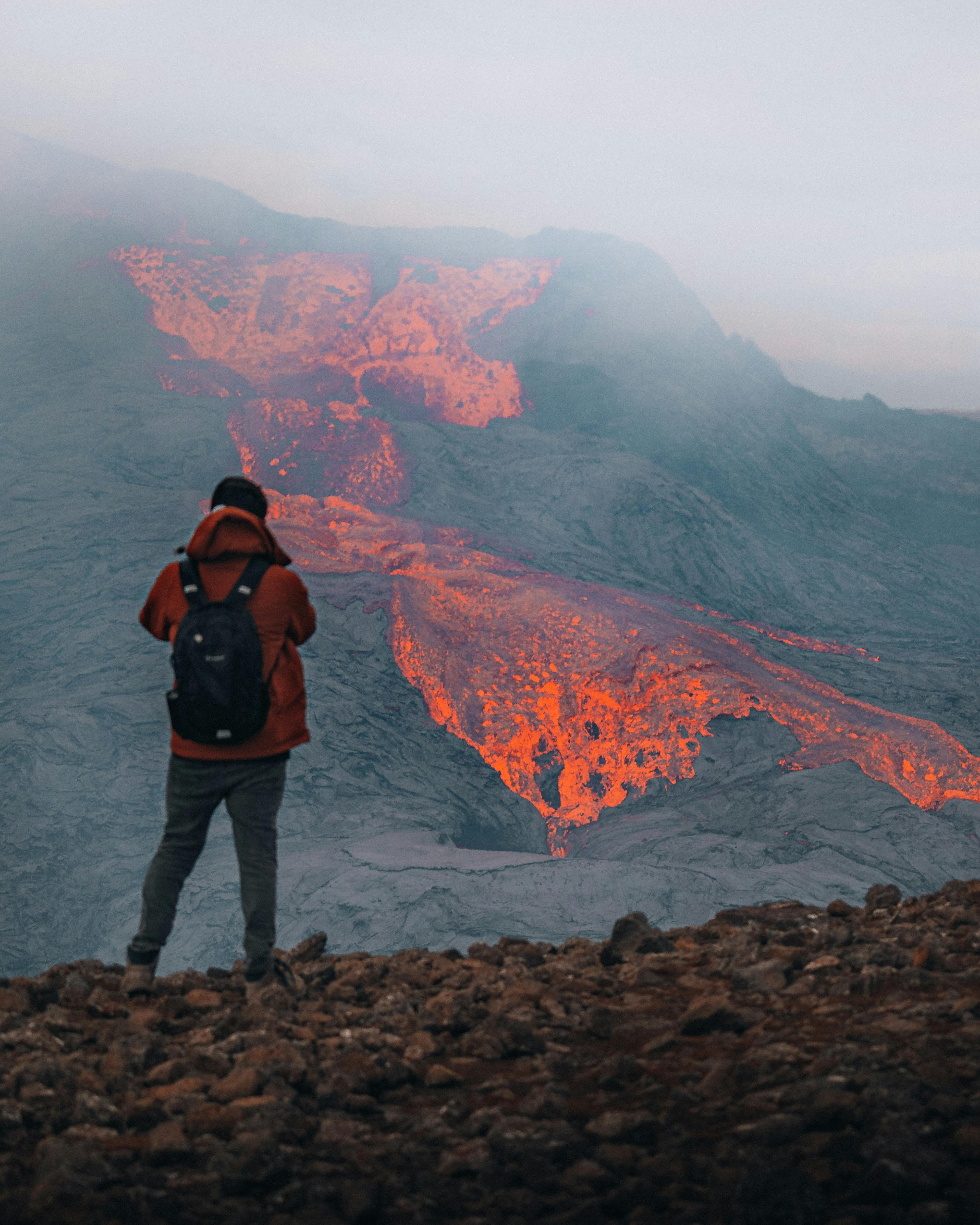 A lone observer clad in a red jacket stands transfixed by the raw beauty of an active lava flow at Fagradalsfjall, a stark contrast of fiery orange against the cooling blues of twilight.