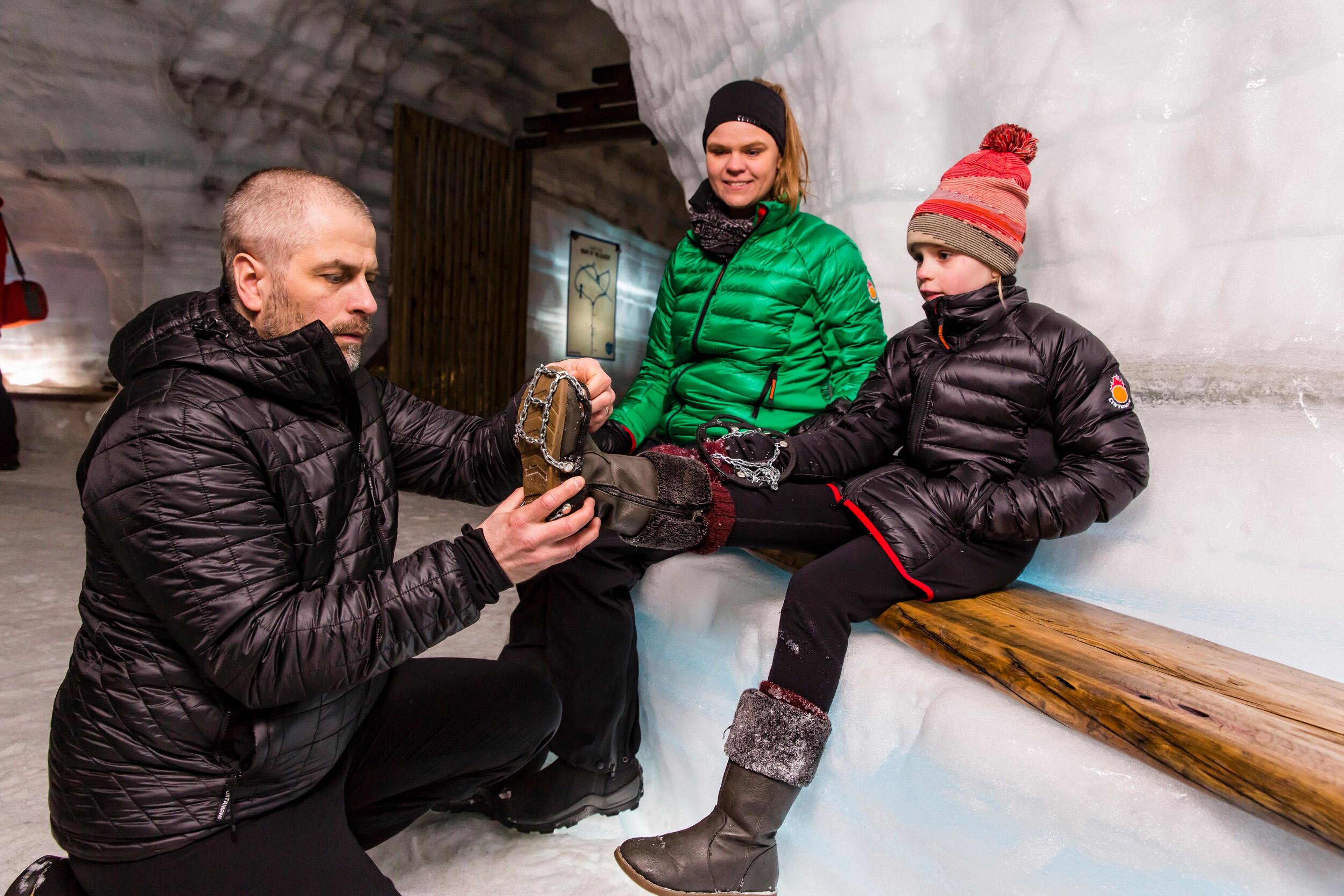 A man adjusts the boot crampons of a child sitting on a wooden bench inside the Langjökull ice cave, with a woman in a green jacket smiling in the background.