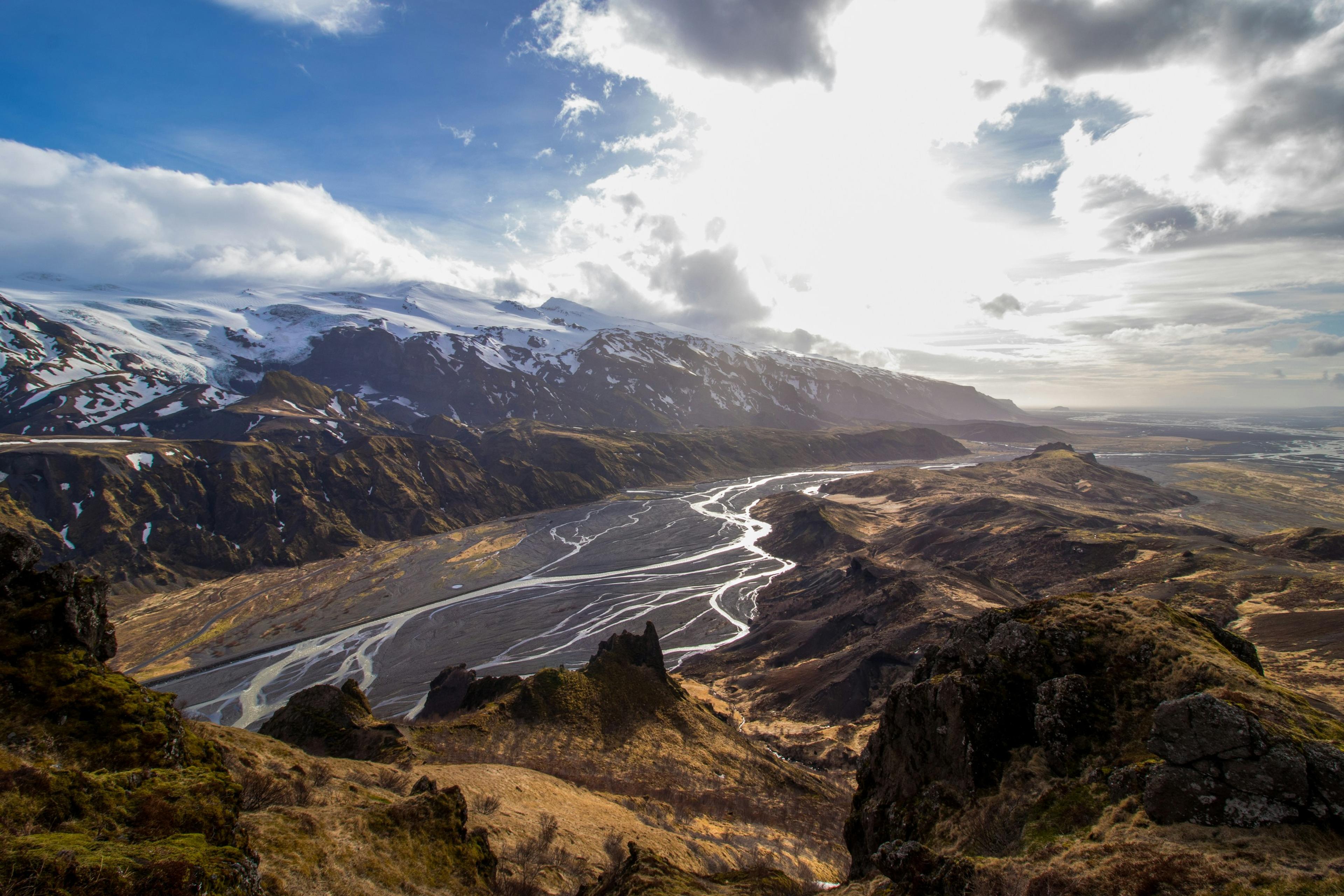 Aerial view of Icelandic Highlands with winding rivers, rugged terrain, and snow-capped mountains under a dramatic sky.