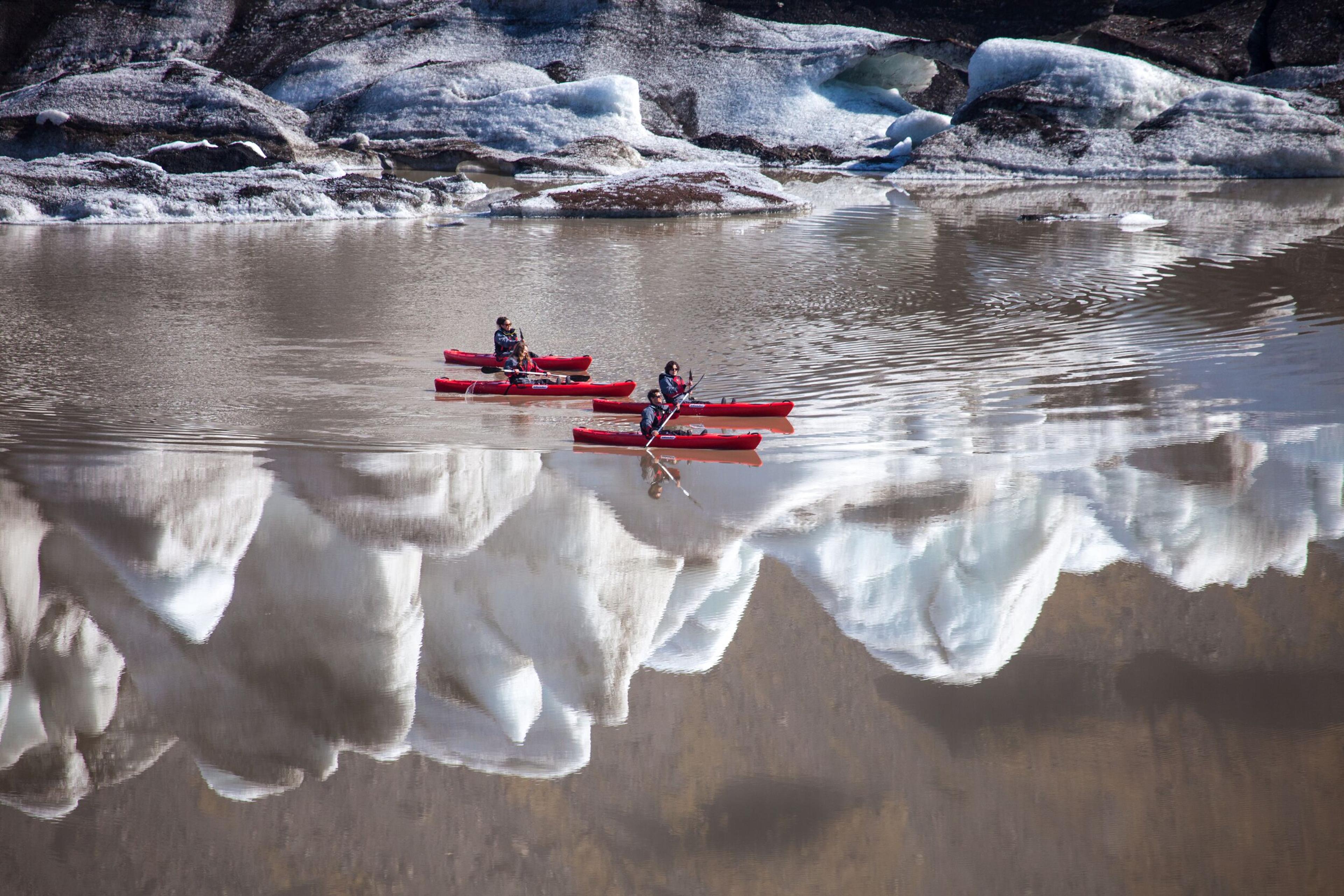 Four kayakers gracefully paddling across a glacier lagoon, with the icebergs beautifully mirrored in the tranquil waters.