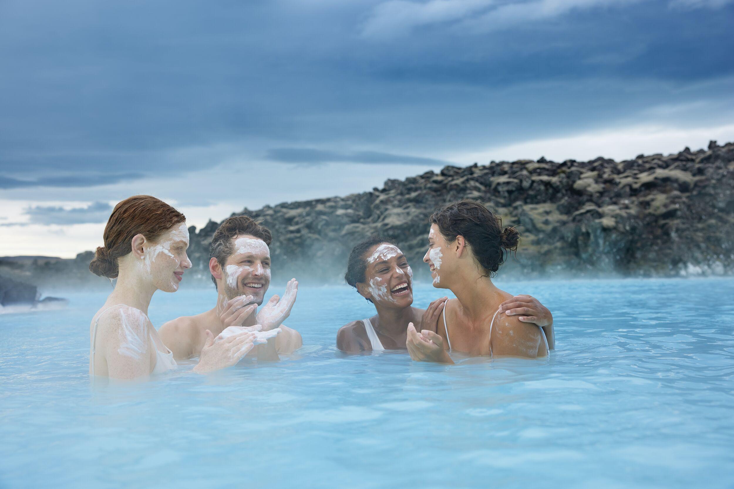 Four smiling people in a milky blue thermal pool, applying white mud to their faces and shoulders.