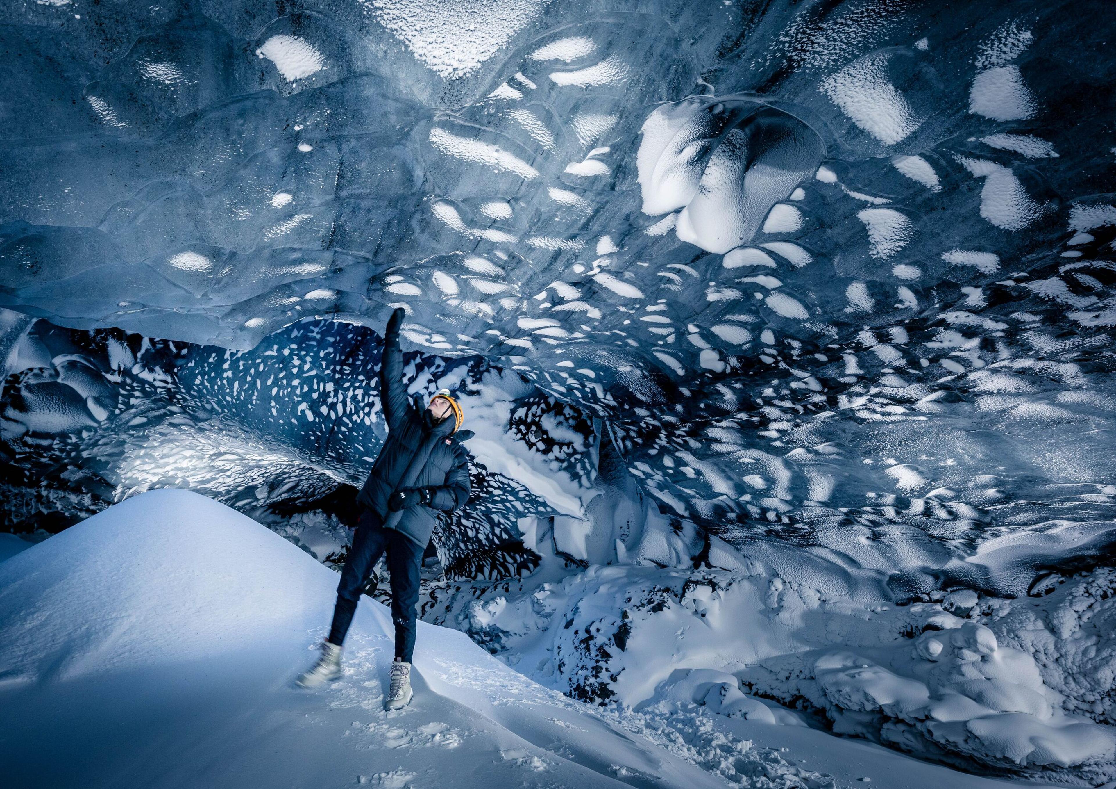A person with raised arms stands at the entrance of a magnificent ice cave, expressing a sense of triumph or awe. The cave's interior is a frozen wonderland, with snow-covered formations that create an otherworldly landscape.