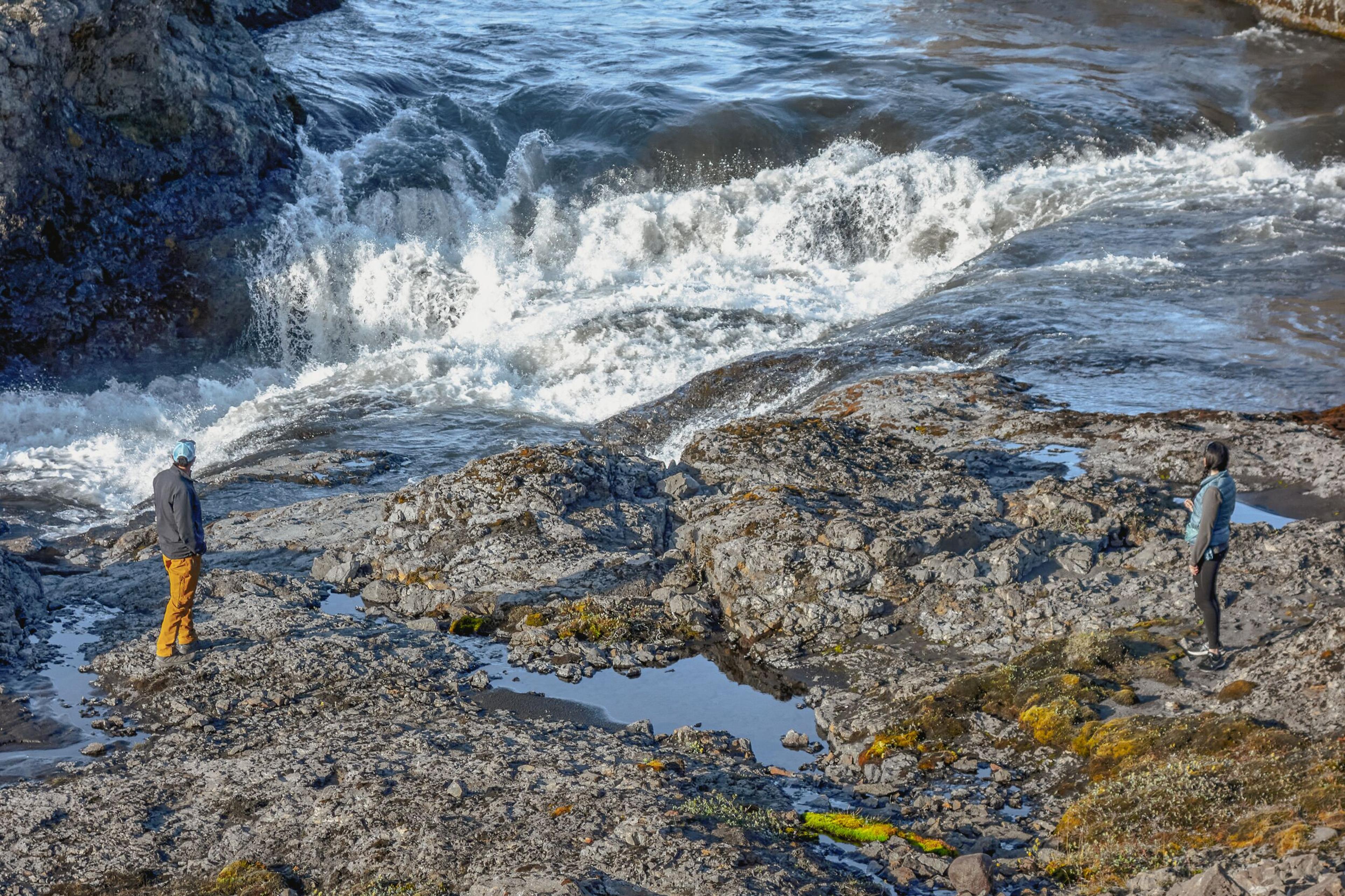 Two people stand on a rocky bank looking at a powerful waterfall.