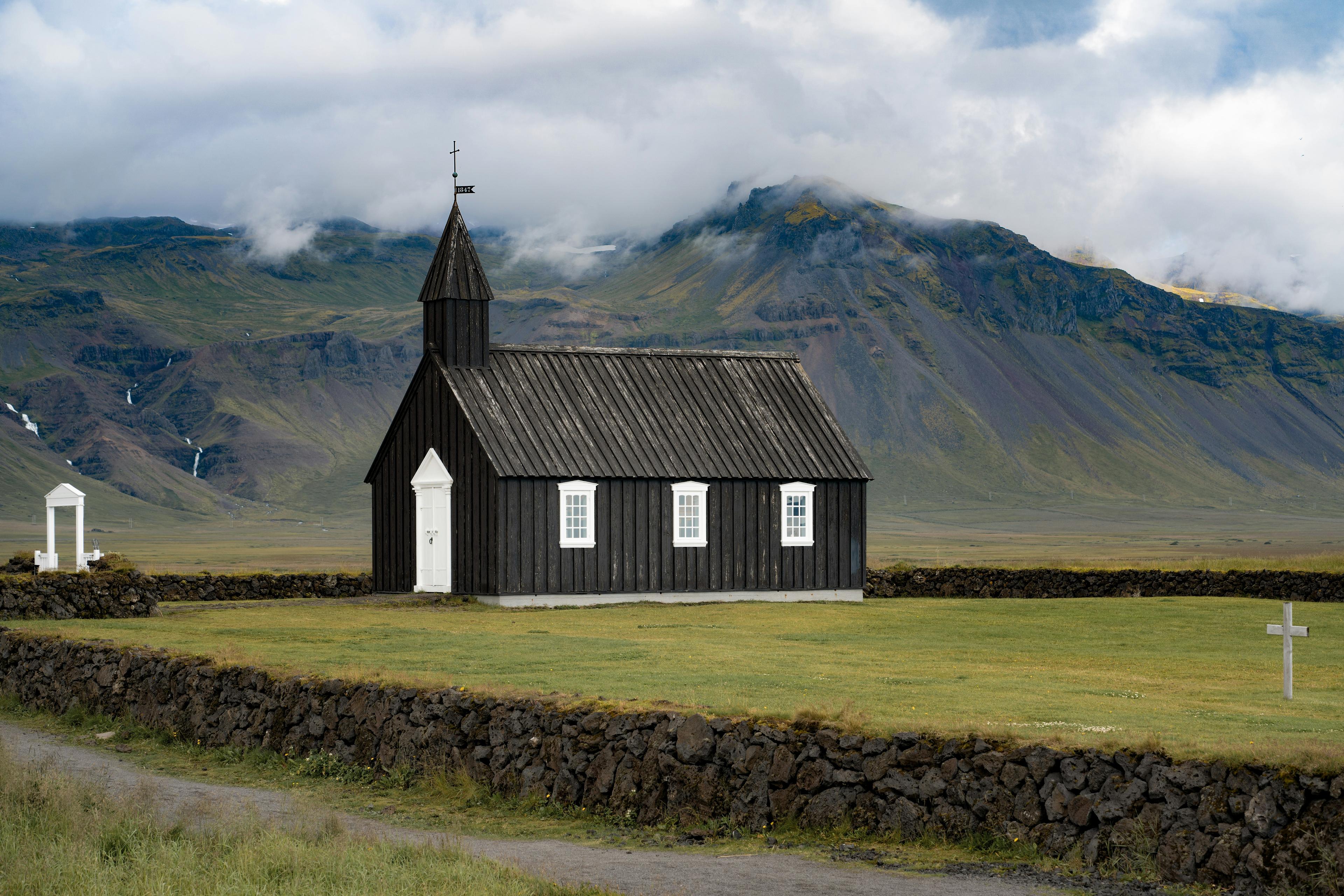 The iconic Búðakirkja, a small black wooden church in Iceland, stands alone against a dramatic mountain backdrop, surrounded by a low stone wall and open grassy fields under a cloudy sky.