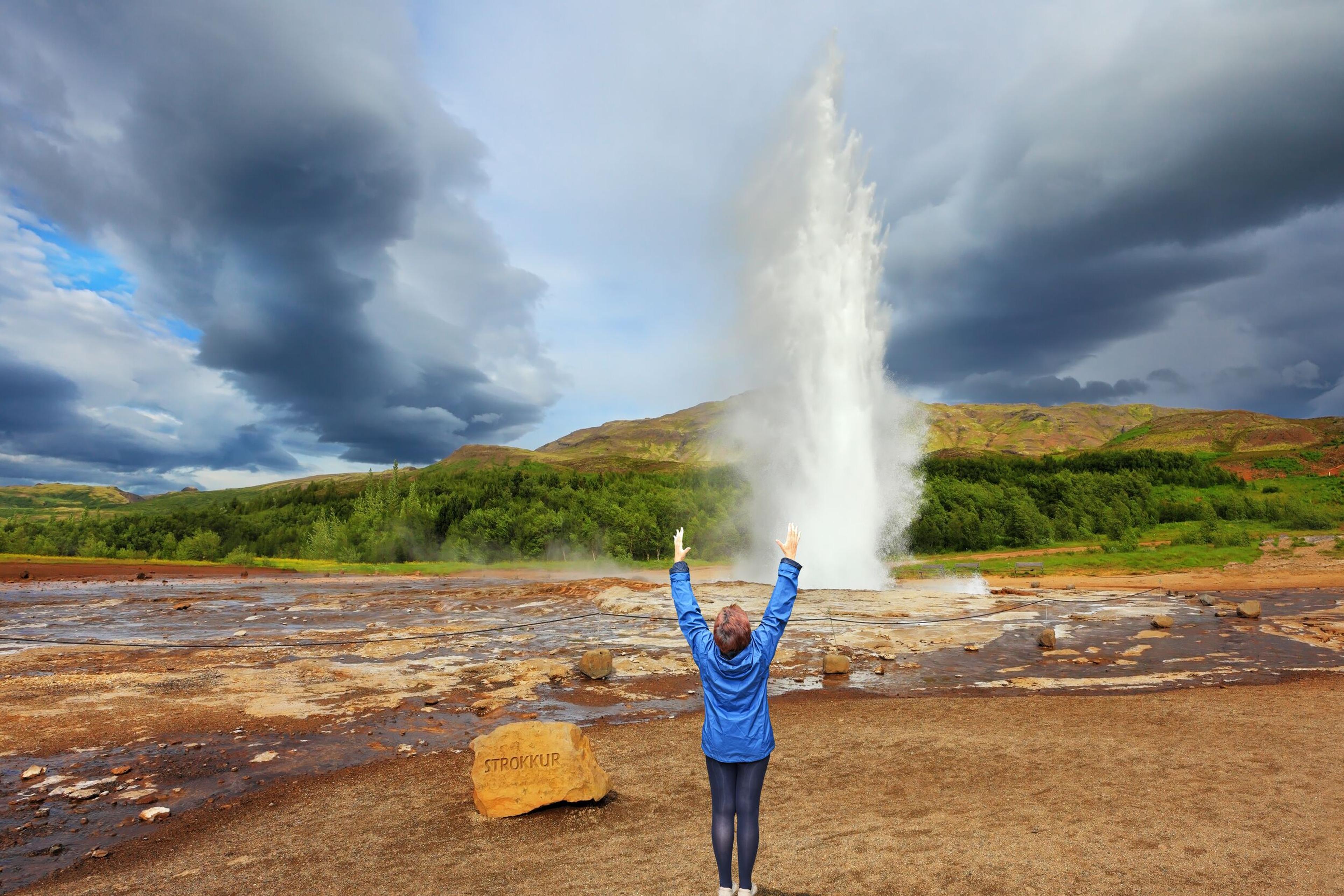 Child in blue jacket with arms raised stands in front of Strokkur geyser erupting at the Golden Circle, Iceland. Dramatic clouds loom overhead, and the ground is marked with geothermal mineral deposits.