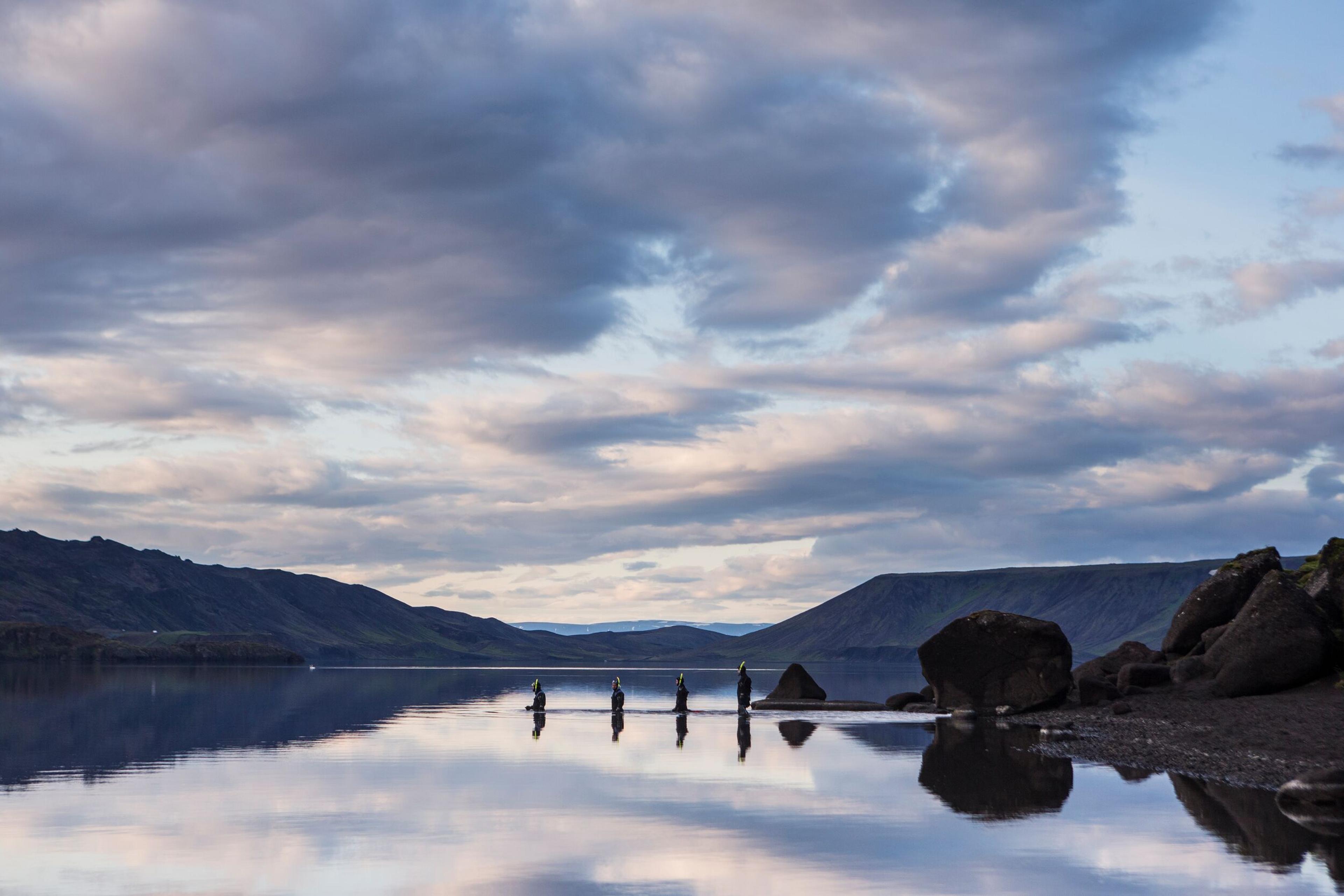 Five people walk in a line across a calm lake, reflecting the cloudy sky and distant hills.