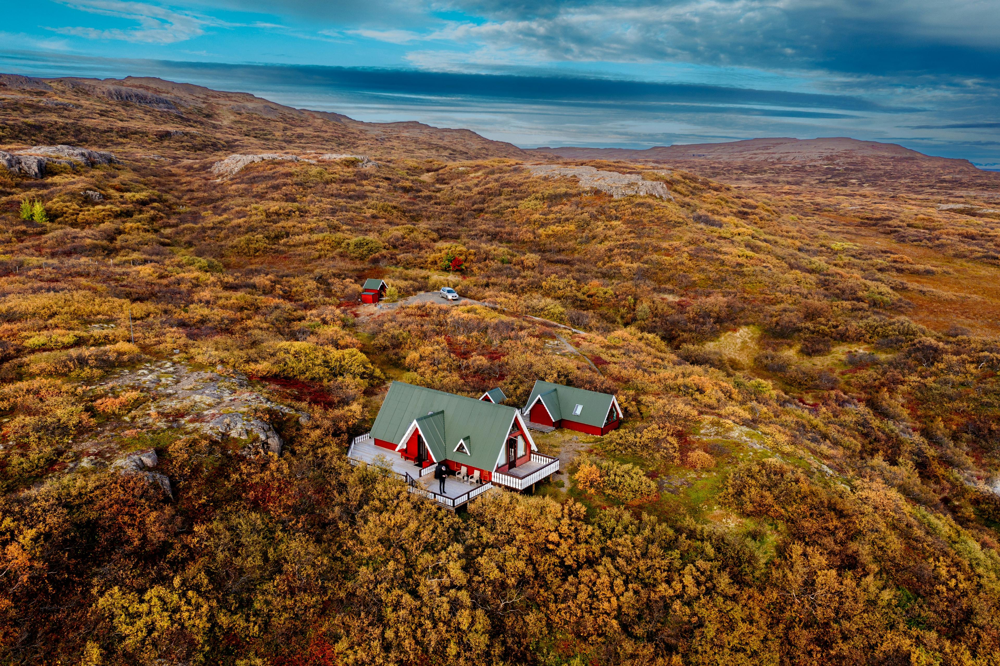 A cluster of red-roofed cabins with green accents nestled in the autumnal landscape of the Borgarfjörður region, surrounded by vibrant fall foliage and rugged Icelandic terrain under a dramatic sky.