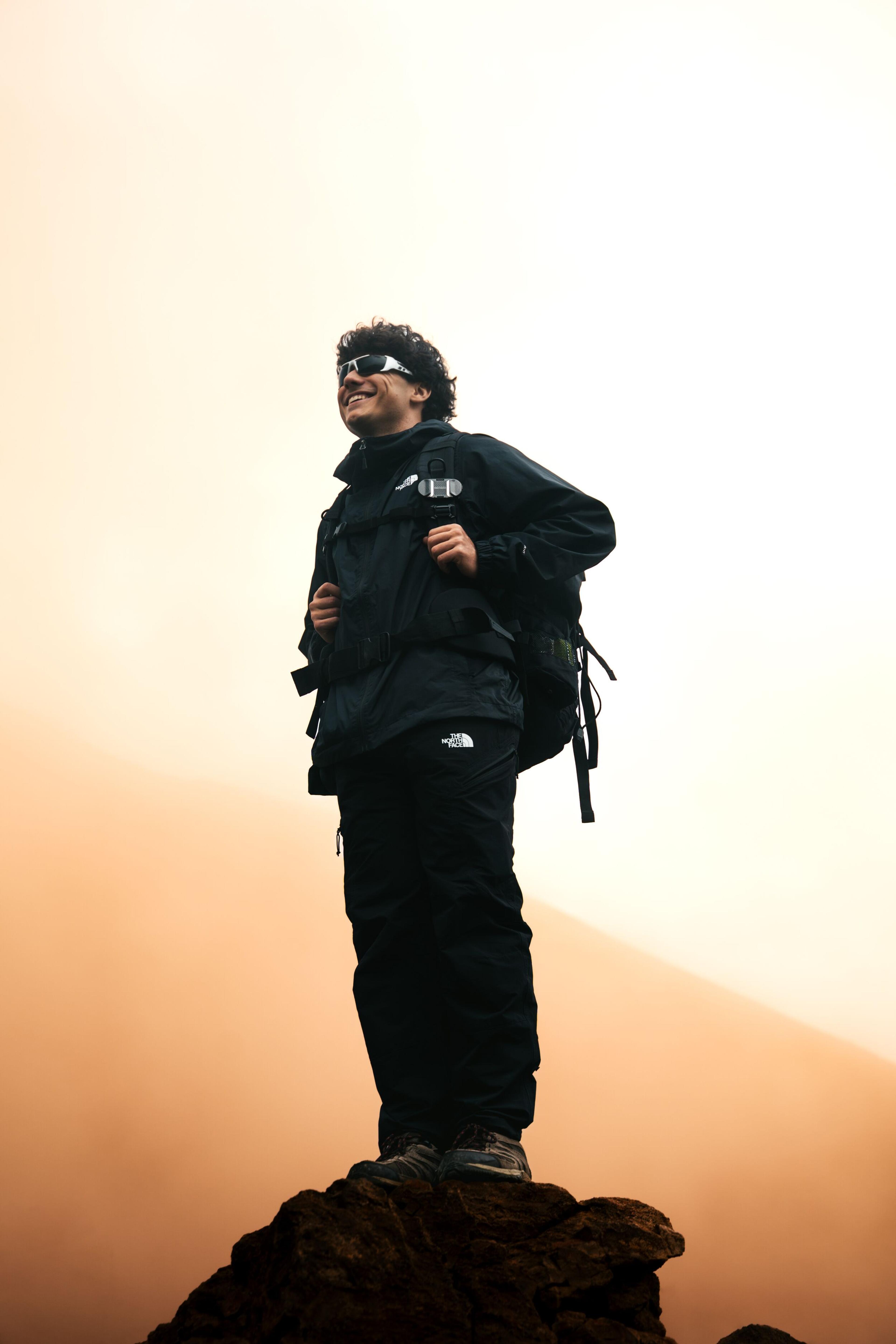 A smiling man in hiking gear and sunglasses stands on a rocky peak, looking up at an orange sky.