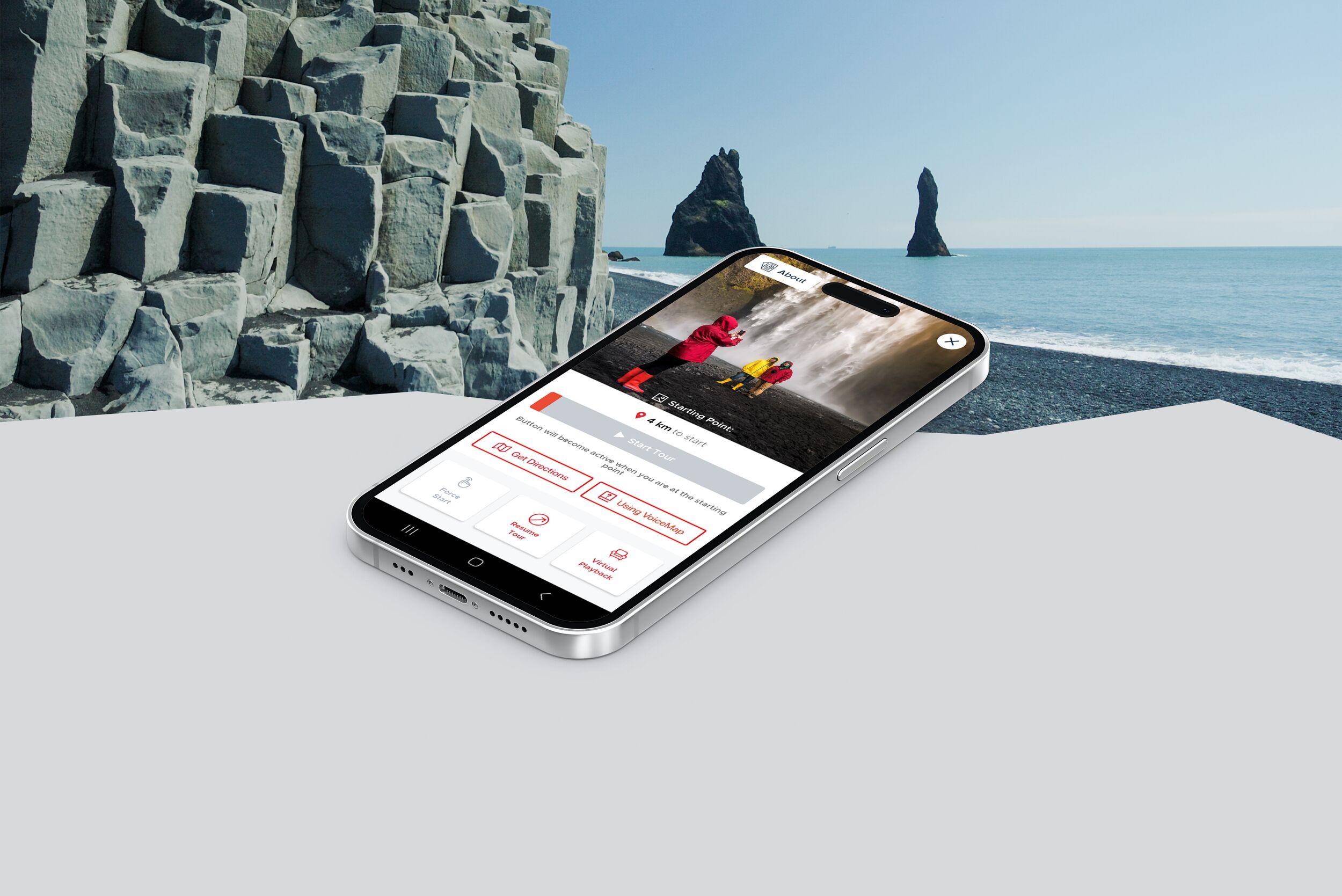 A smartphone displaying VioceMap audio guide app rests on a white surface with the screen showing a location with red markers, set against a striking background featuring the black sands and towering basalt sea stacks of Reynisfjara beach in Iceland.