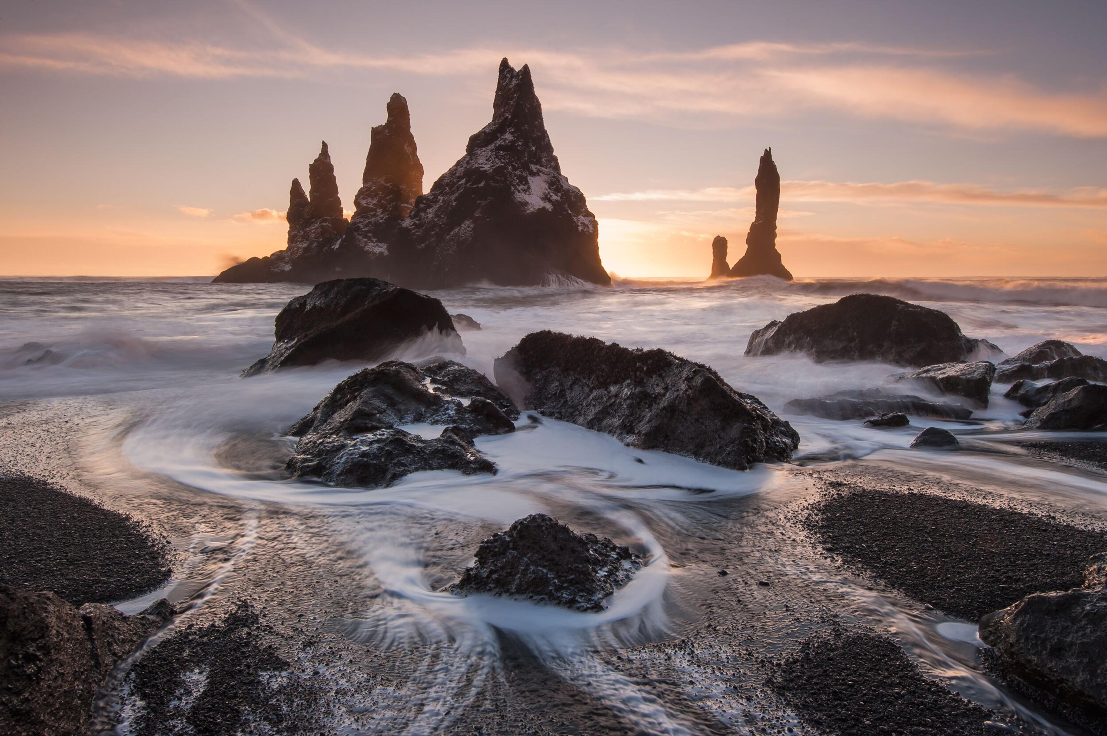 Seascape at sunset with waves swirling around jagged rock formations protruding from the sea, set against a vibrant sky.