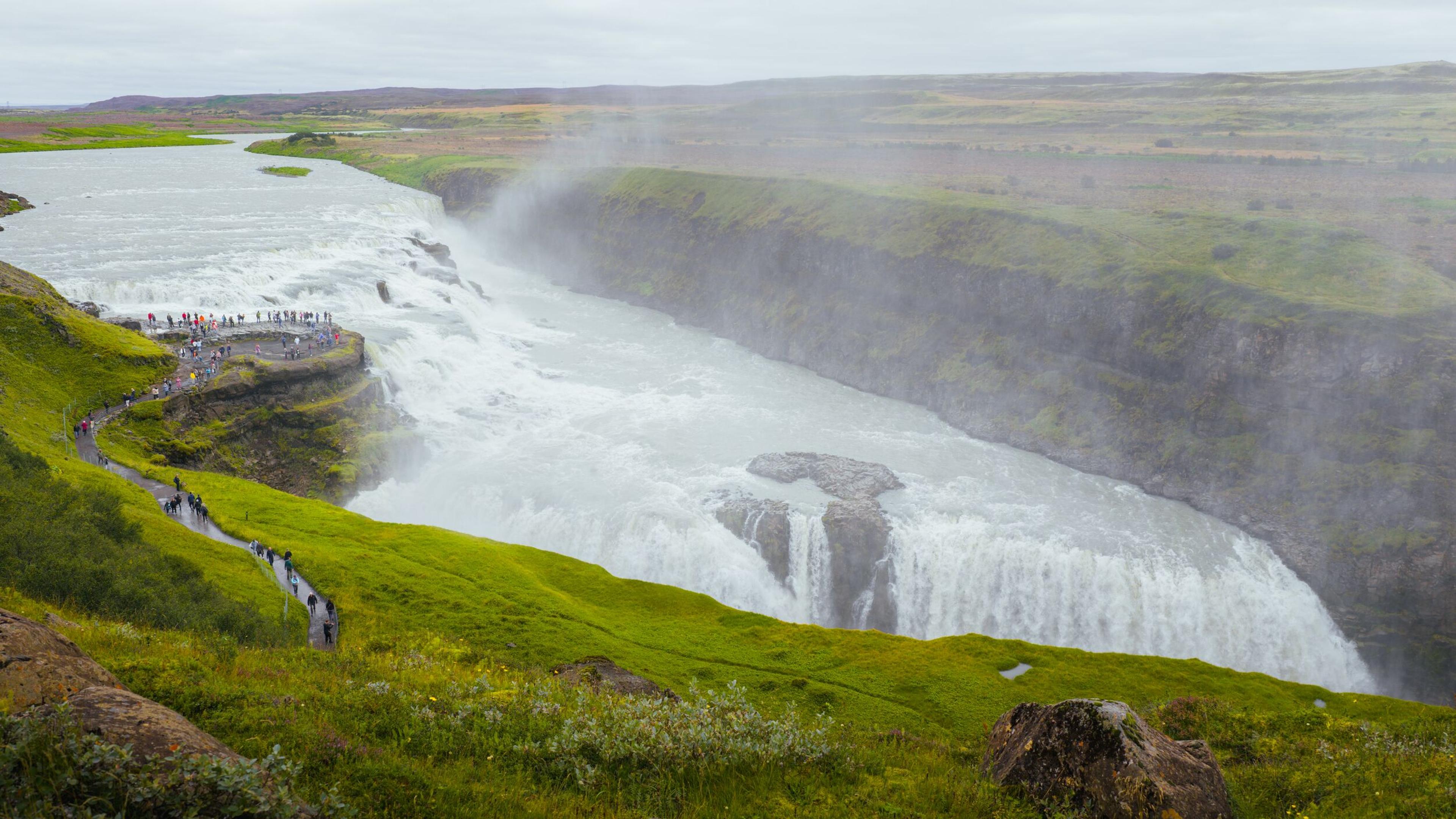 Gullfoss waterfall crashes into a rocky canyon, with a viewing platform crowded with tourists and a winding path on the left, amidst green hills.