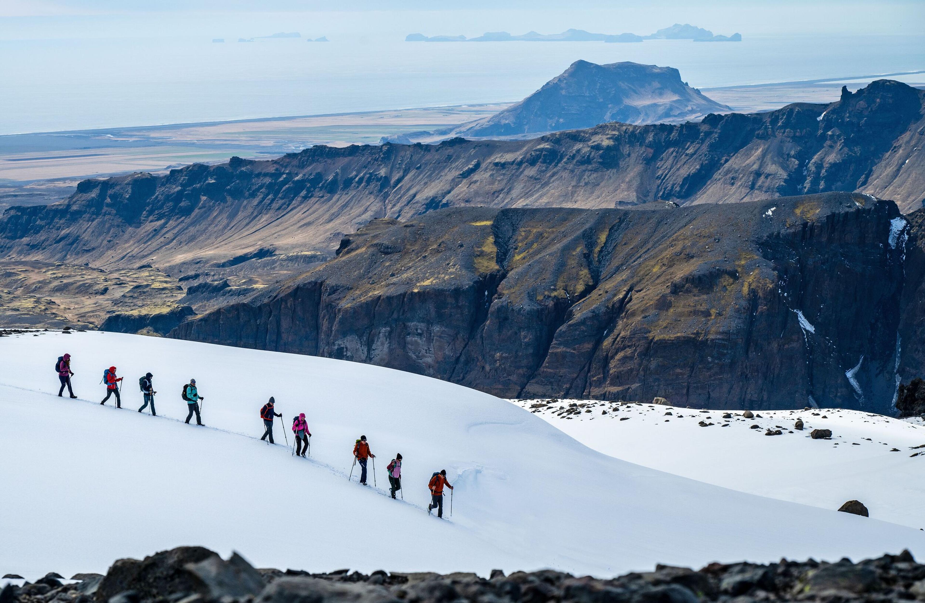 A group of hikers with backpacks walking in a line across a snowy ridge with stunning mountain cliffs and a distant view of the ocean in the background. 