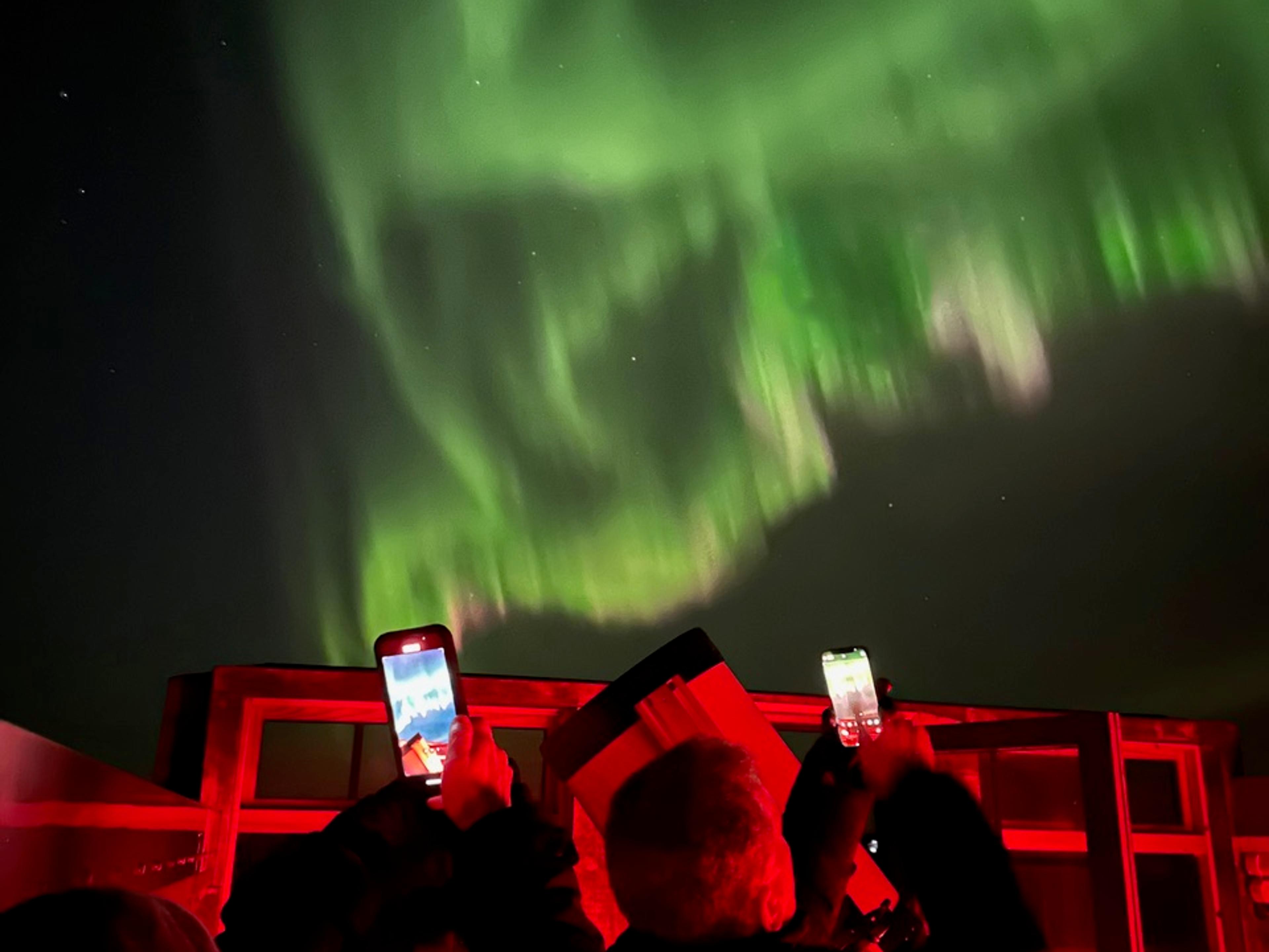 Two people capture the vibrant green Northern Lights on their phones, standing under red-lit surroundings at night.