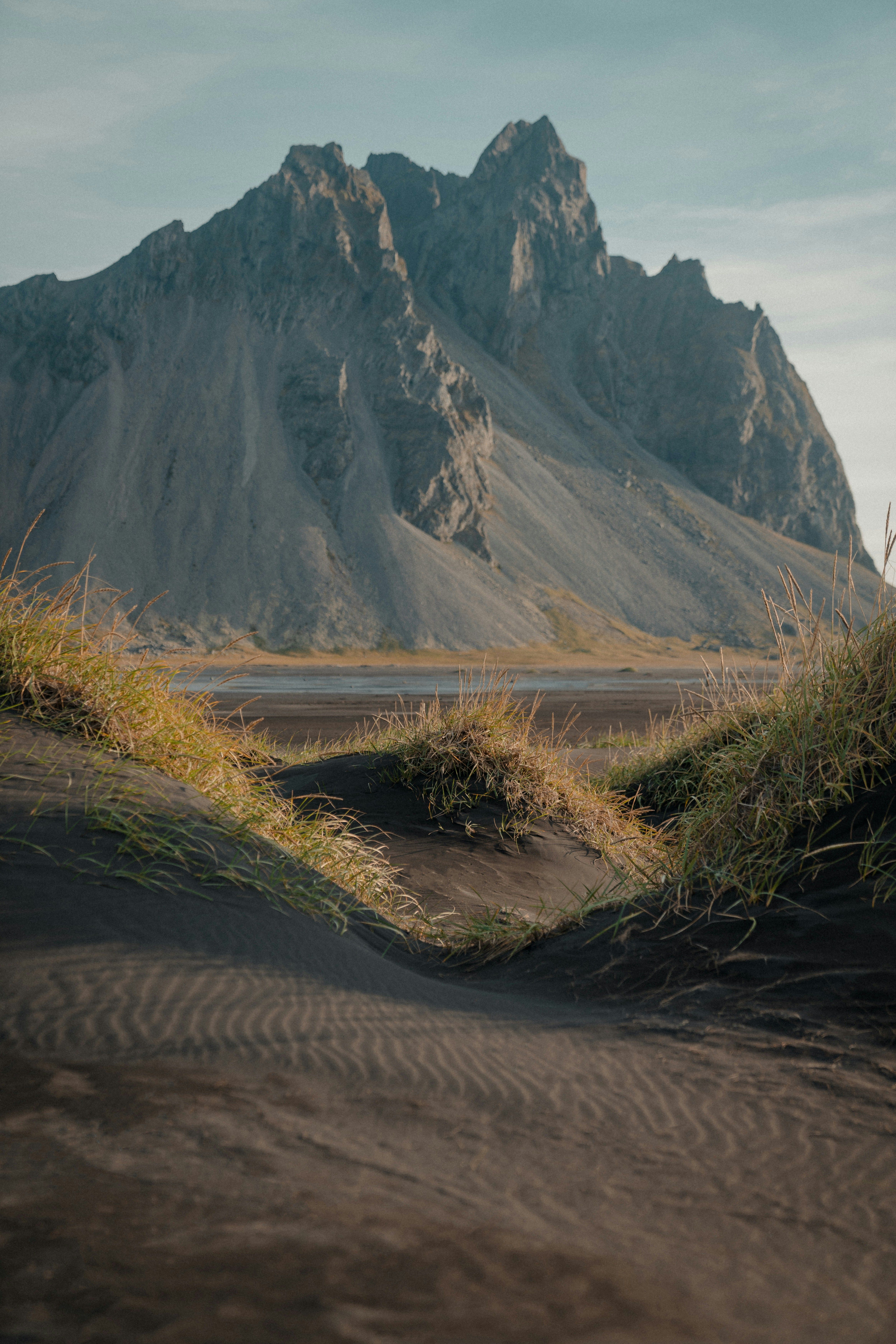 This image showcases the dramatic Stokksnes mountain in Iceland, viewed through black sand dunes topped with golden grass. The jagged peaks rise sharply against the soft blue sky, while the rippled patterns in the sand add depth and texture to the foreground.