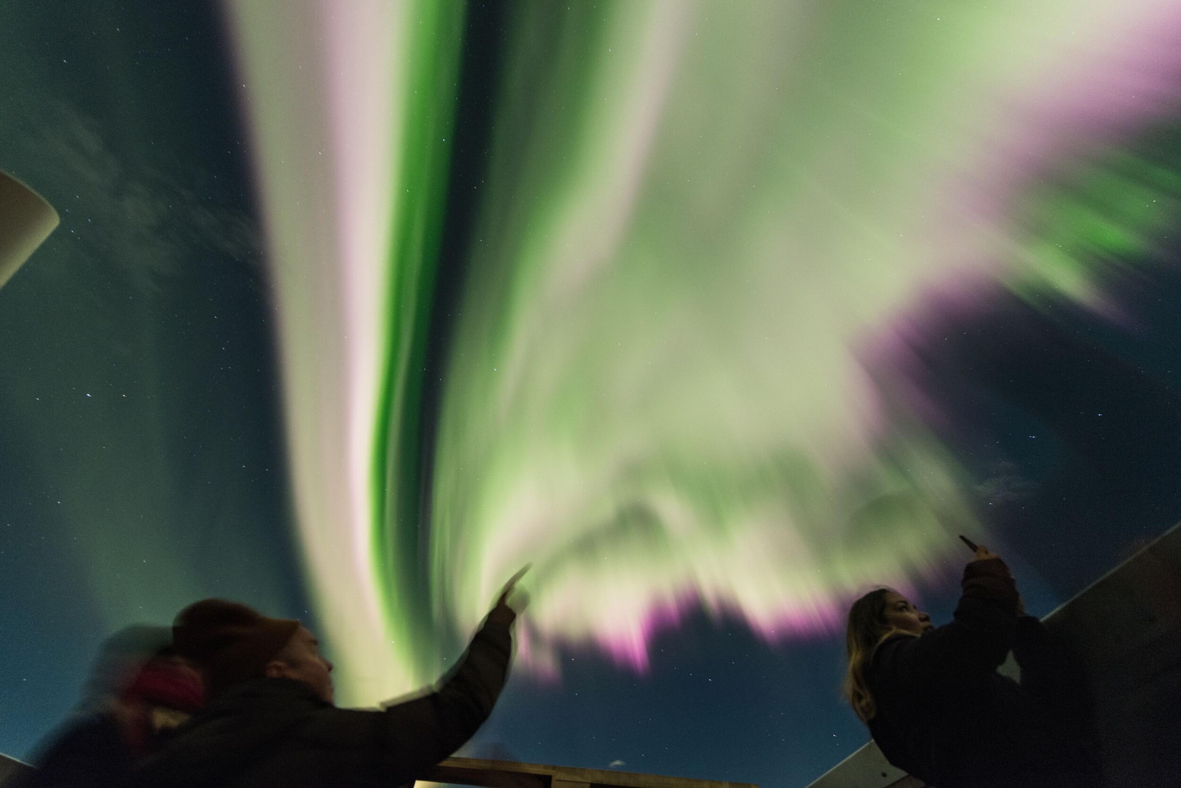 Two people point towards vibrant green and purple Northern Lights as they streak across the dark night sky.