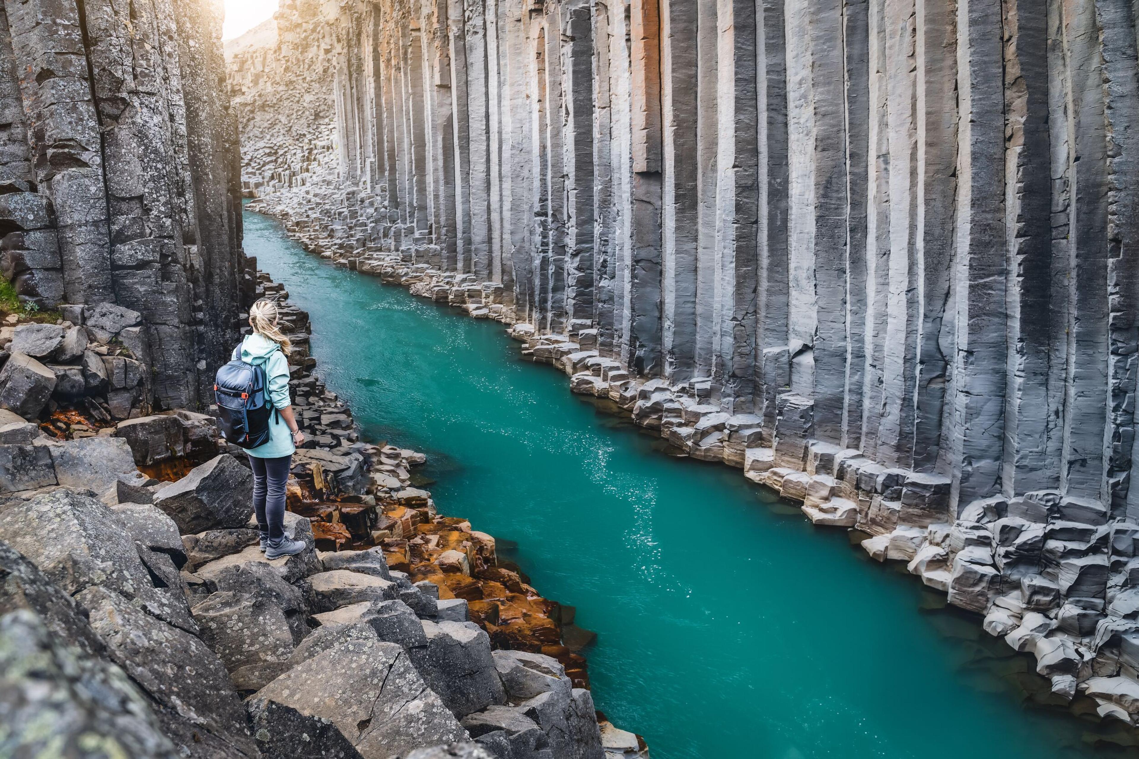 A hiker stands on a rocky path beside a vibrant blue river in a canyon formed by tall basalt columns.
