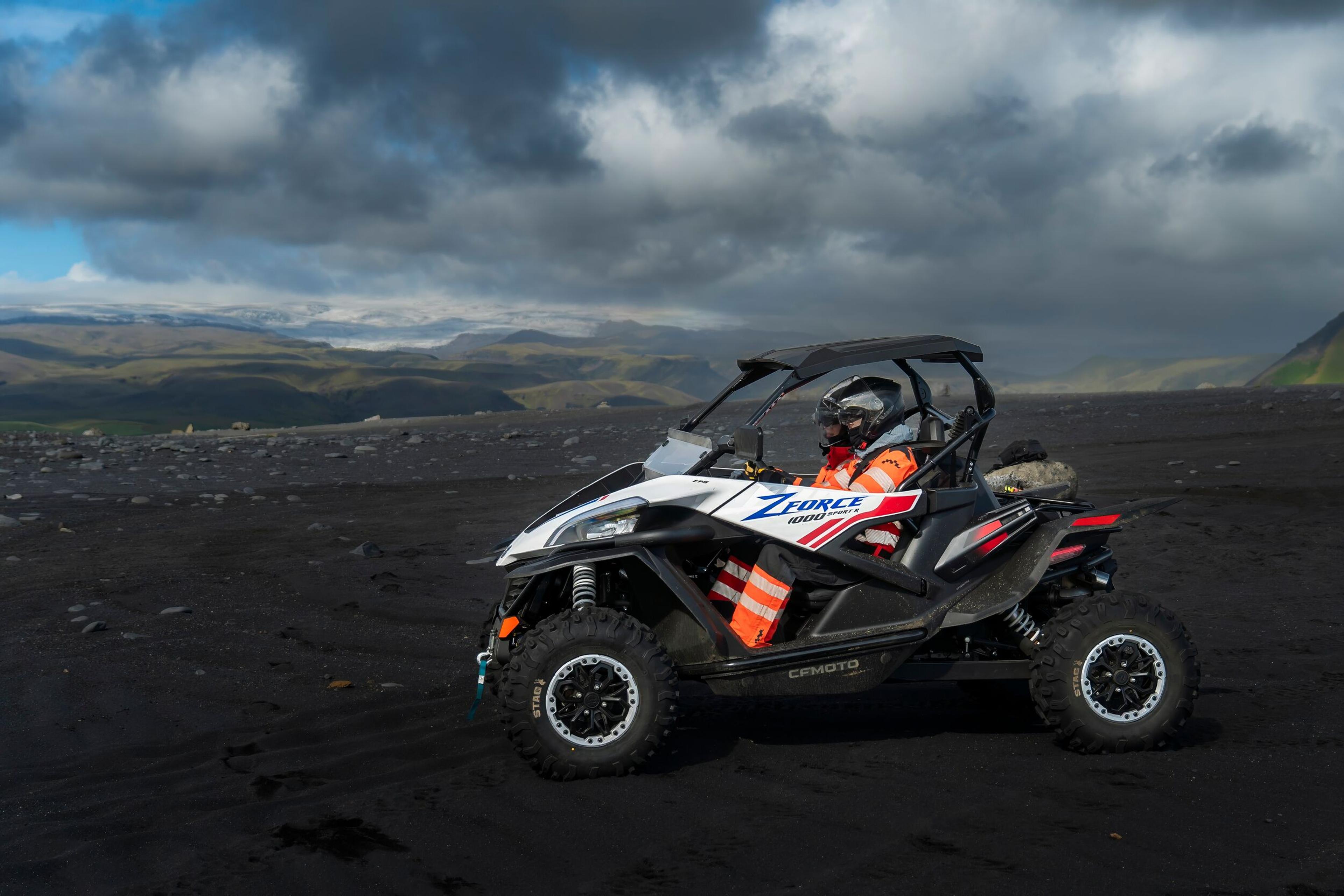 A single off-road buggy with orange and white detailing, situated on a vast black sand beach with a dramatic mountainous landscape in the distance, possibly ready for a buggy adventure in Iceland.