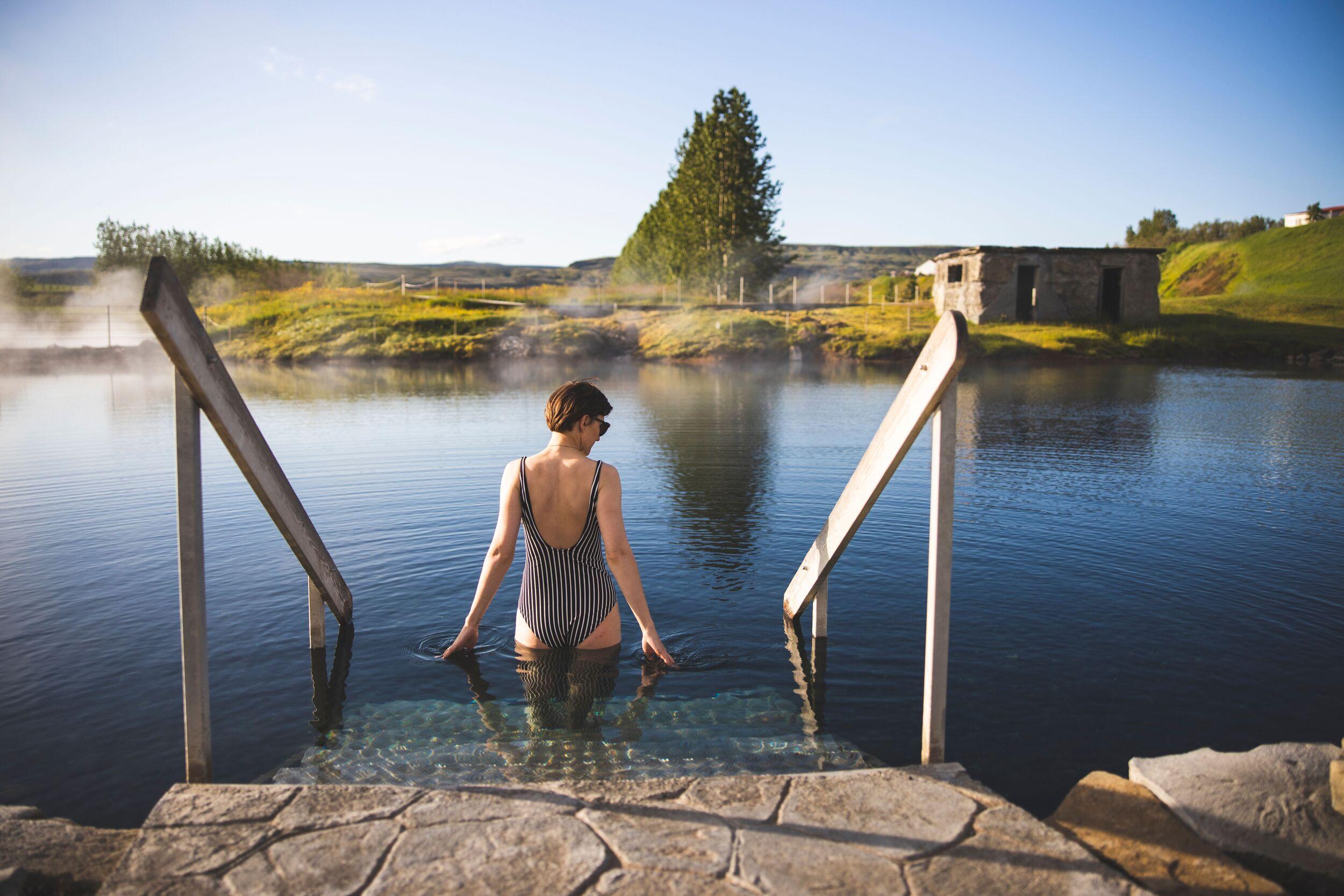 A woman in a striped swimsuit stands on steps in a steaming natural pool, surrounded by a green landscape.