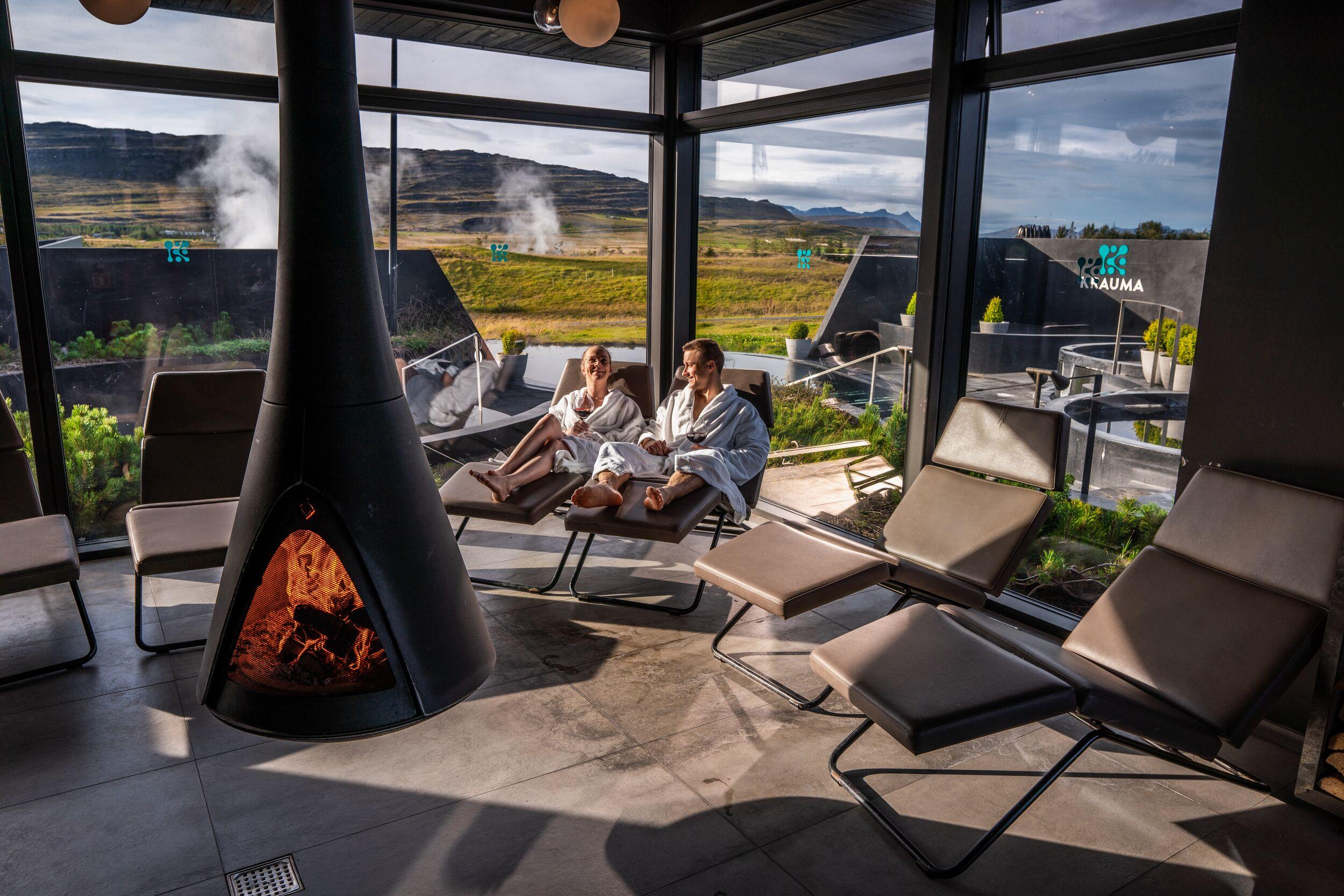 A couple in bathrobes relaxes by a modern fireplace in an indoor spa lounge with large windows overlooking outdoor geothermal pools and a scenic landscape.