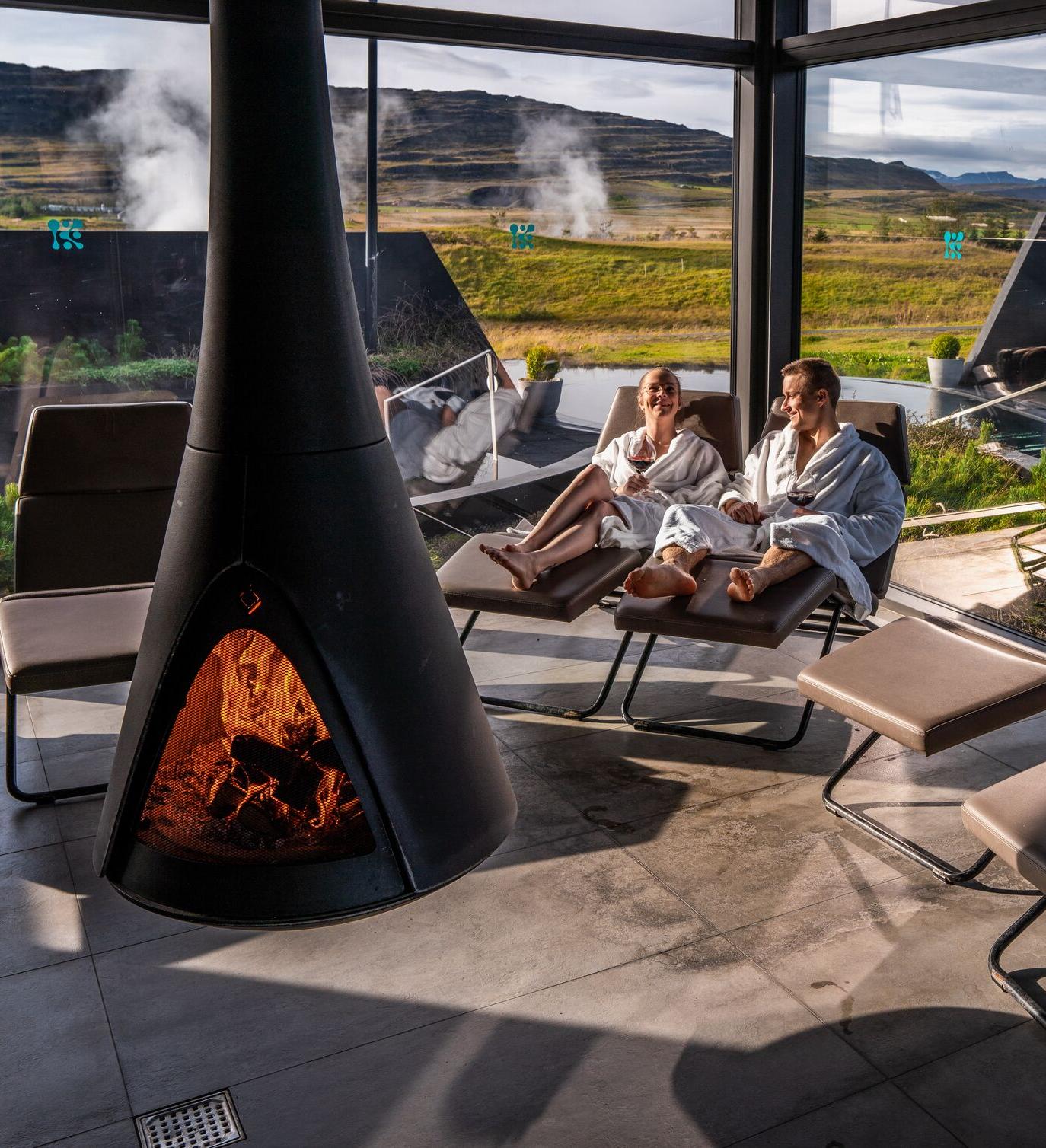 A cozy relaxation room at Krauma Baths features a modern black fireplace with a warm fire, while two guests in white robes lounge on chairs, enjoying the view of geothermal steam rising from the Icelandic landscape through large glass windows.