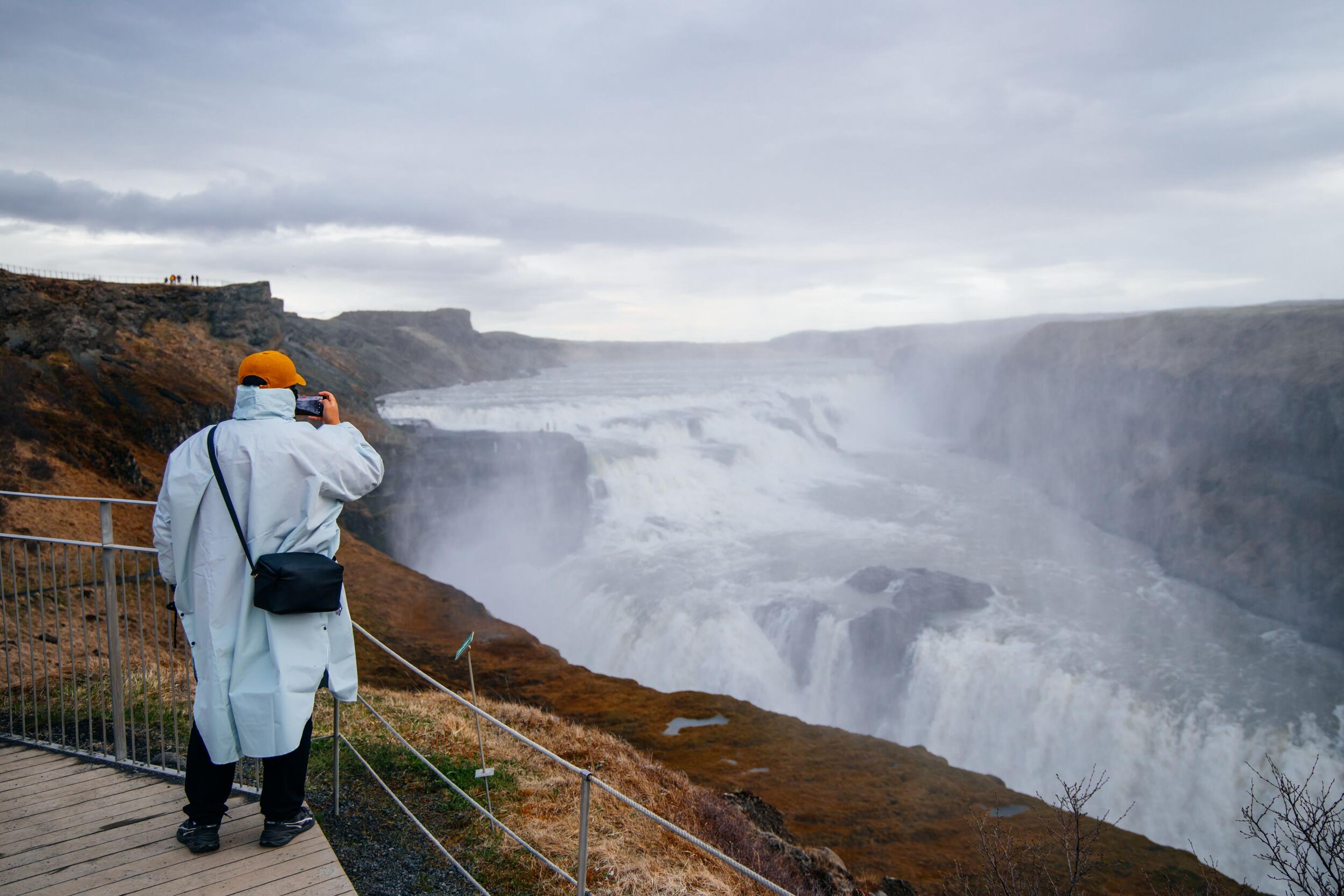 A person in a raincoat and orange hat photographs a large, misty waterfall from a viewpoint.