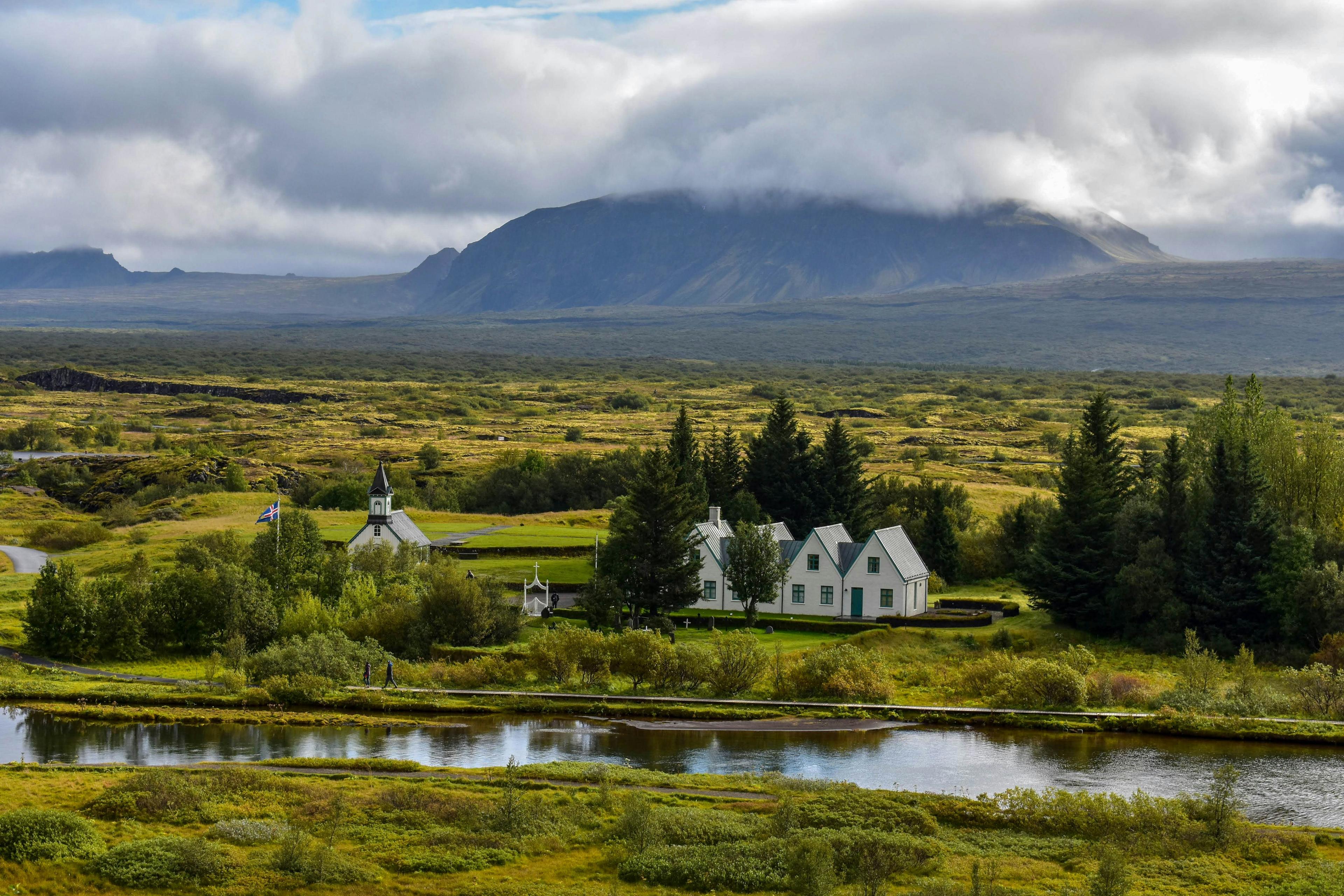 A serene landscape bathed in warm sunlight, featuring a small settlement with traditional buildings near a river, nestled within the vast, open plains and gentle hills in Þingvellir (Thingvellir) National Park in Iceland.