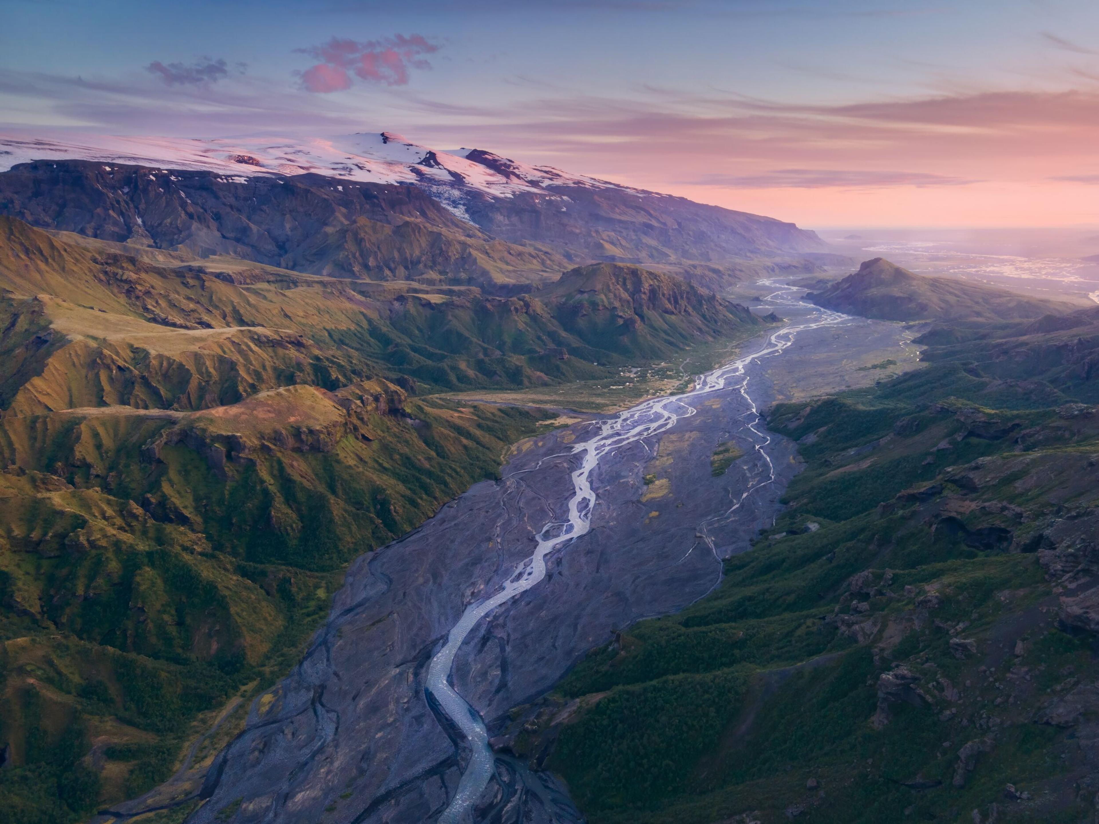 Spectacular river valley flanked by craggy hills, with a distant glacier, all bathed in the soft pink hues of sunset.