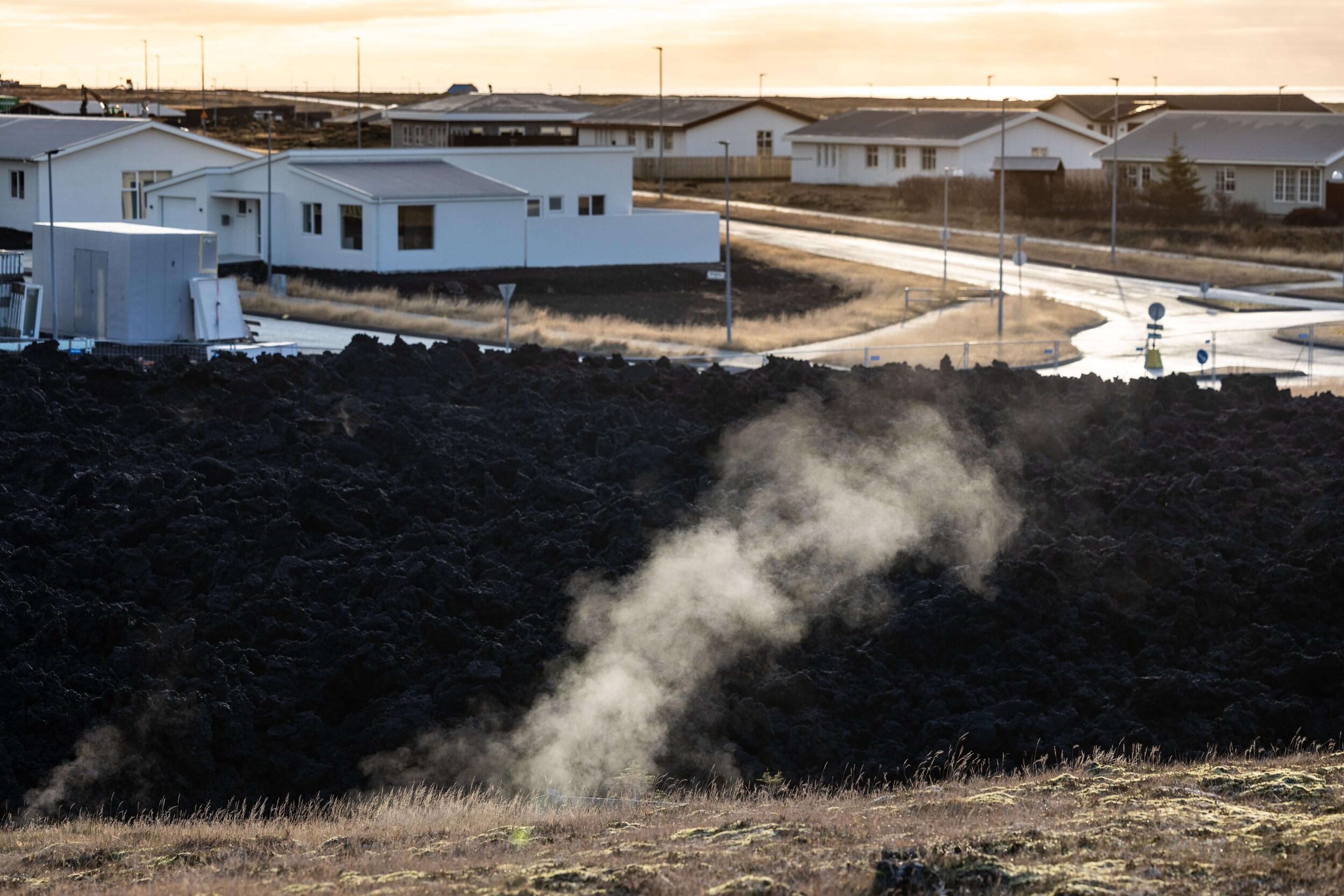 Steam rising from a rugged lava field with a neighborhood of white houses in the background.