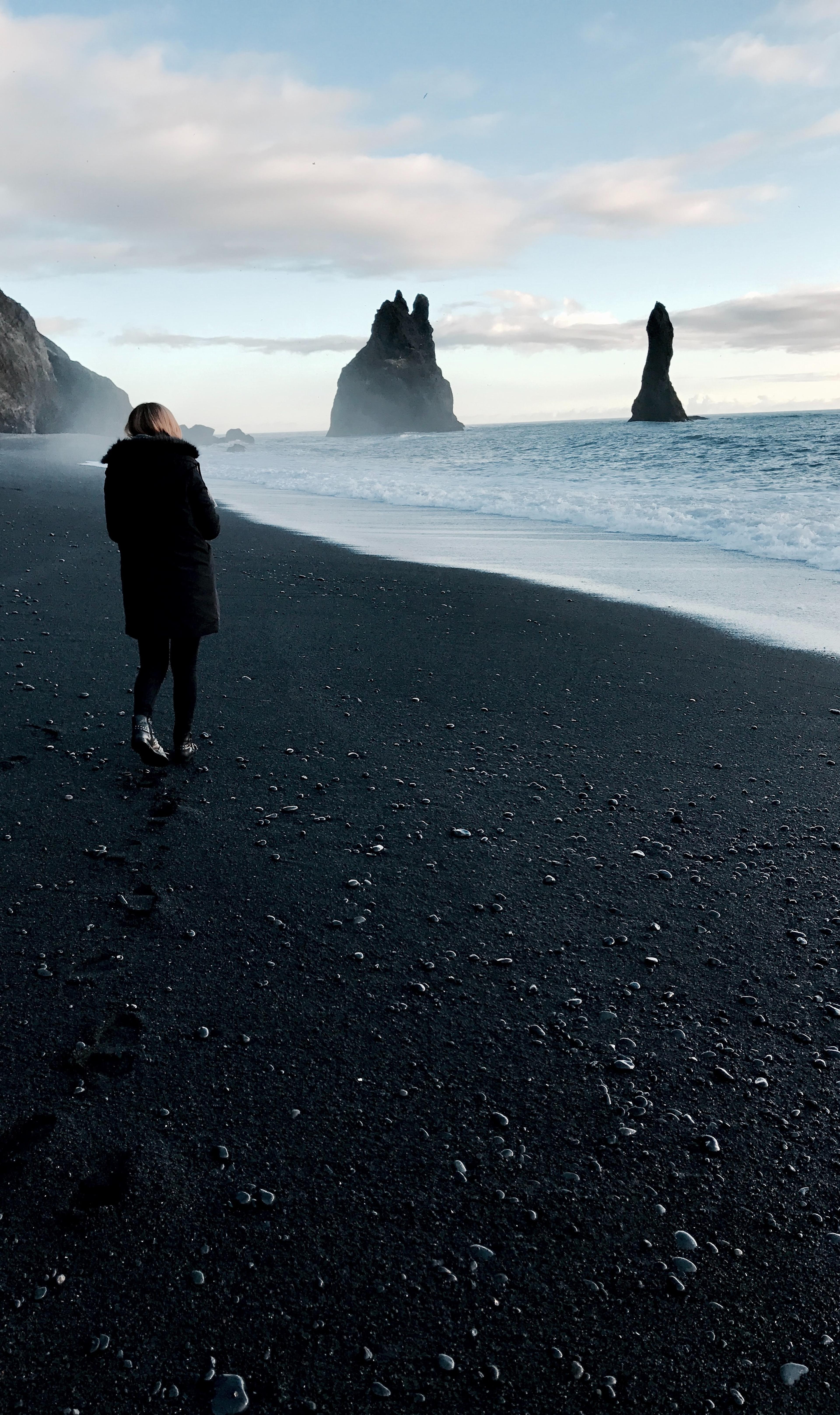 A woman strolling on the black sand beach, wrapped in a voluminous black parka.