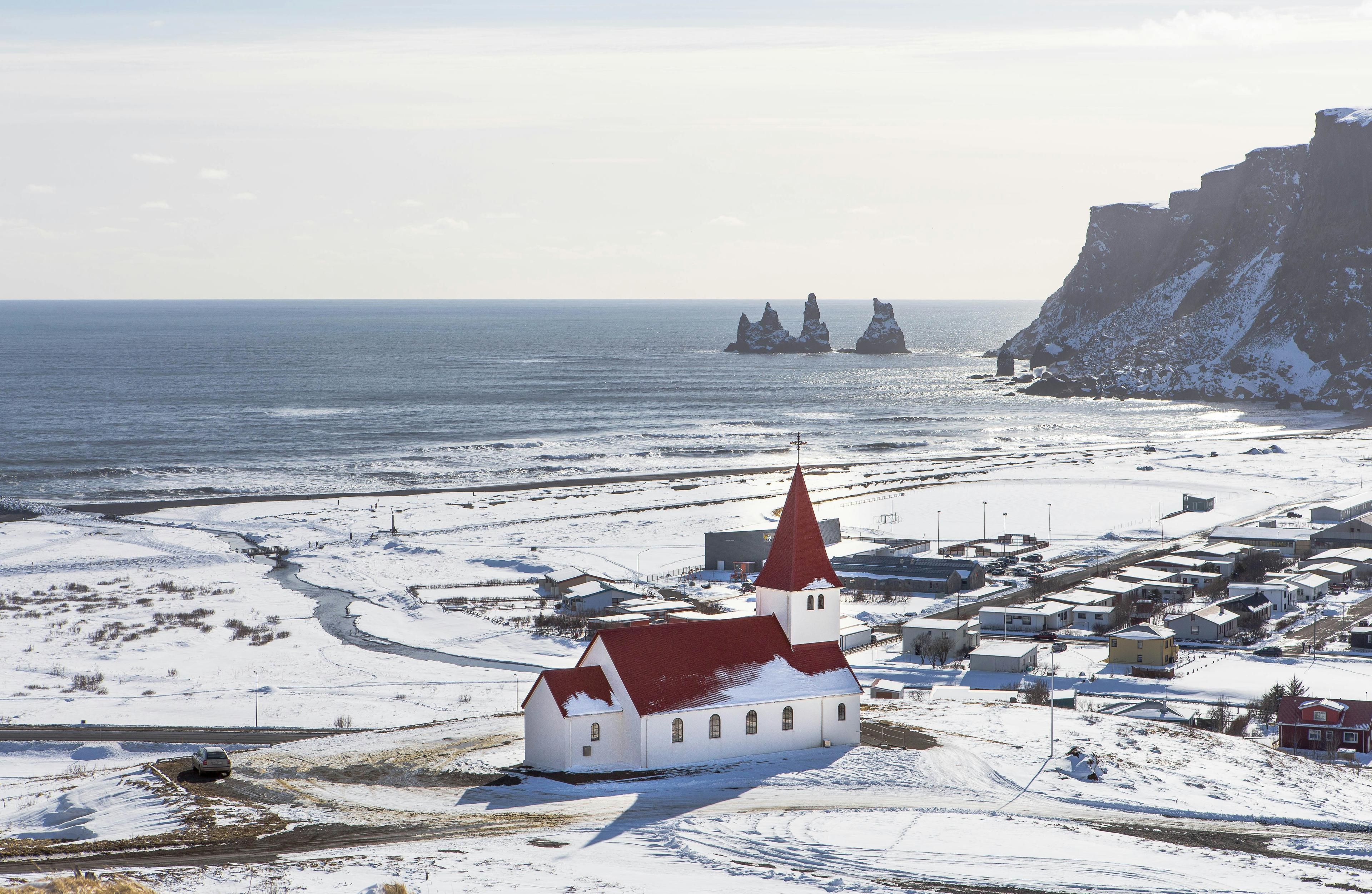  The red-roofed Vík church stands out against snow-covered scenery and ocean views, featuring Reynisdrangar sea stacks on Iceland's South Coast.