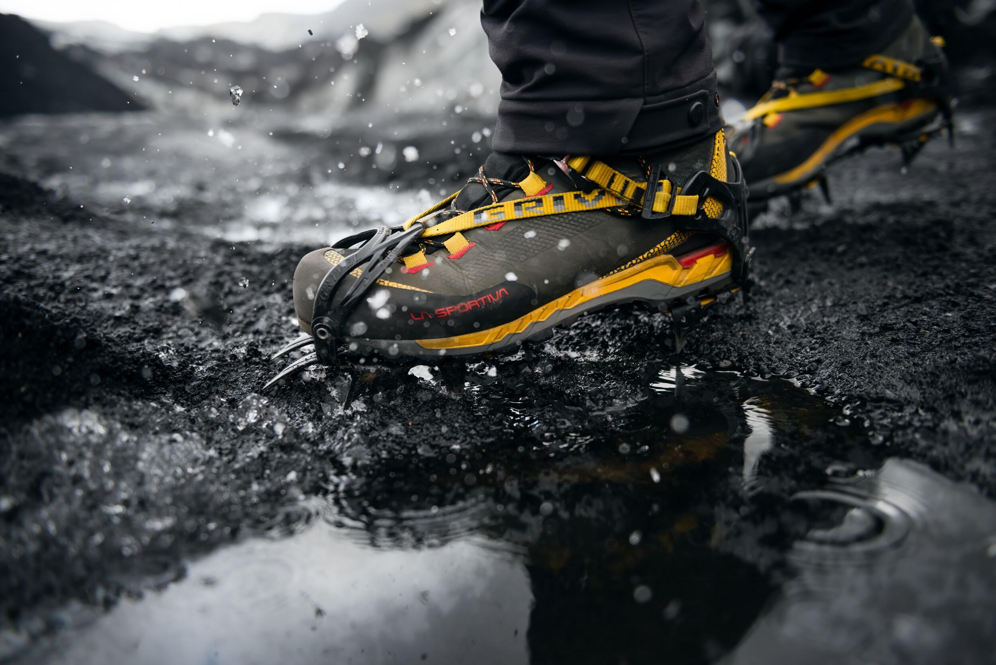 Close-up of a hiker's boots with crampons walking through muddy terrain, splashing water.