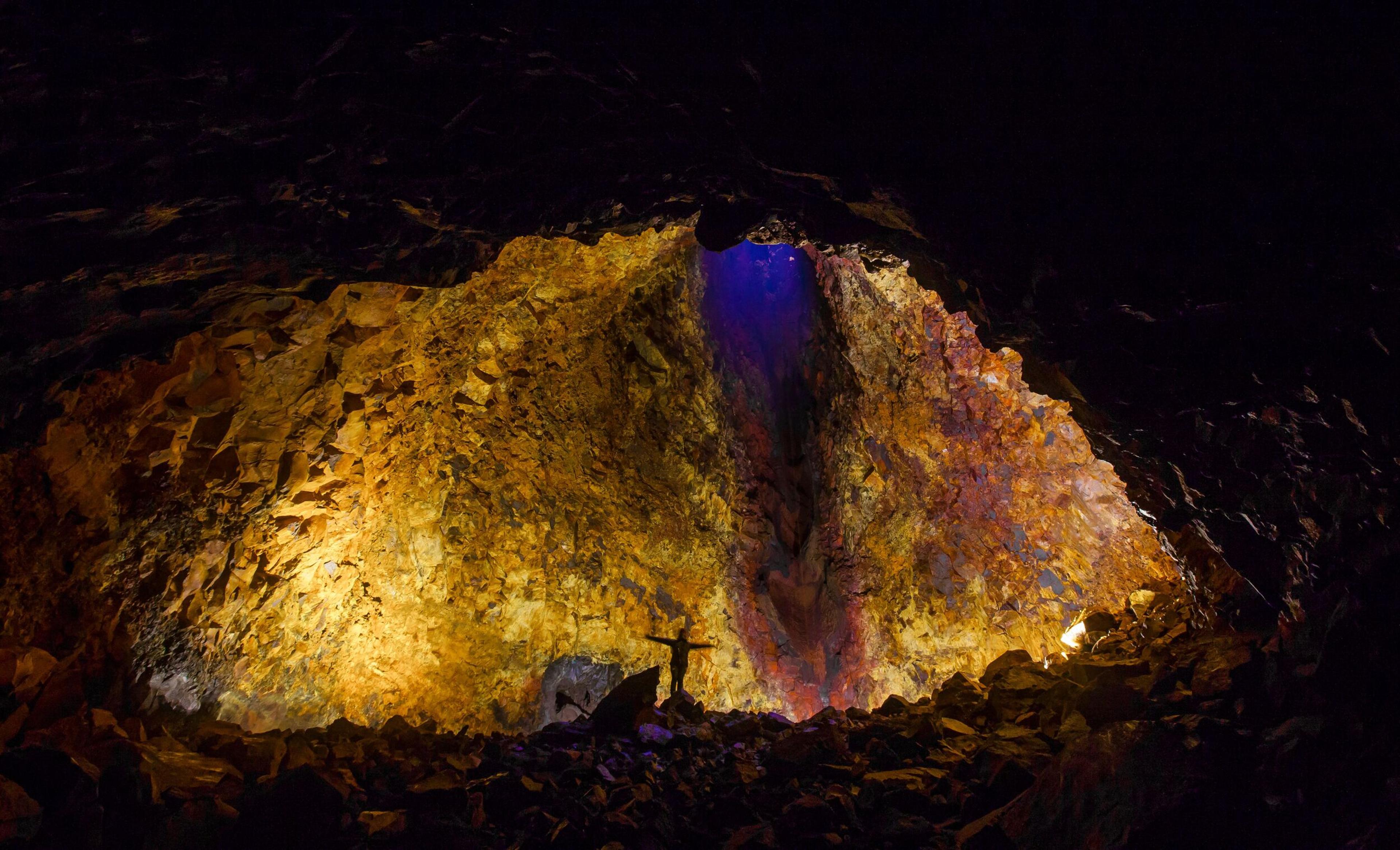 A person with outstretched arms stands in a large, vibrantly lit volcanic cave.