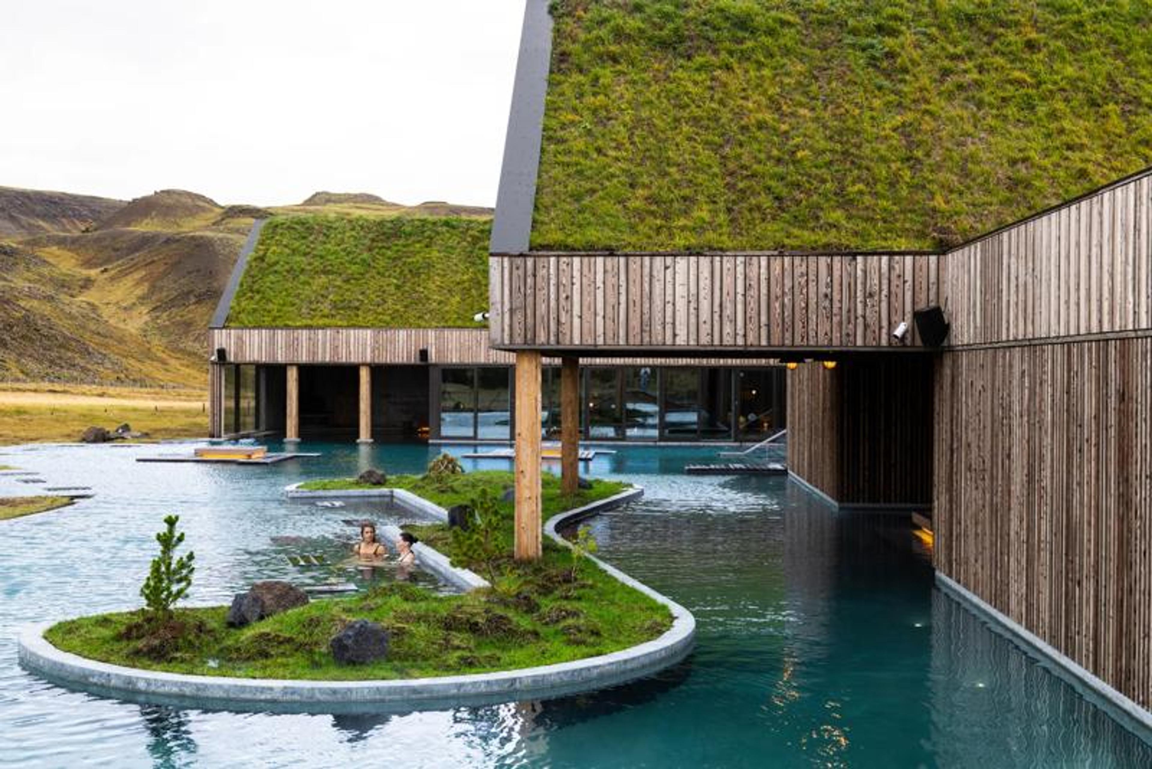 Outdoor geothermal spa with green-roofed buildings, a blue lagoon, and two bathers.