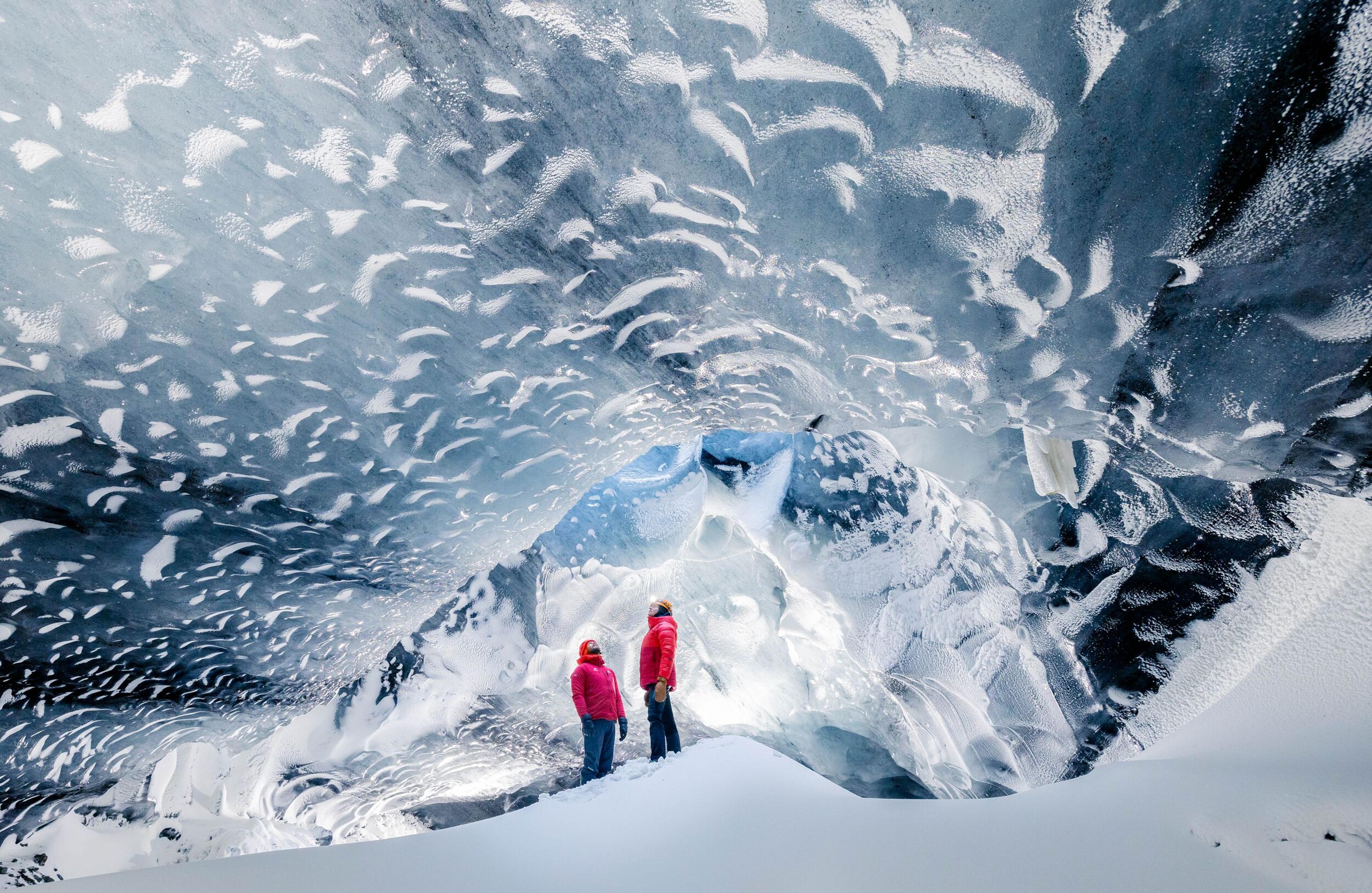 Two people in bright jackets standing at the entrance of a vast ice cave, with one person pointing towards the inside. The cave's inner walls glisten with layers of ice, and the snow on the ground enhances the cave's ethereal beauty.